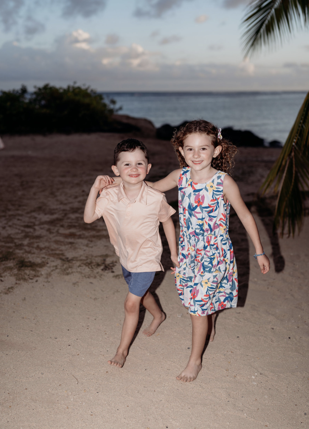 Two children, a boy and a girl, playing together on a sandy beach during sunset, smiling and running barefoot, with ocean and a cloudy sky in the background.