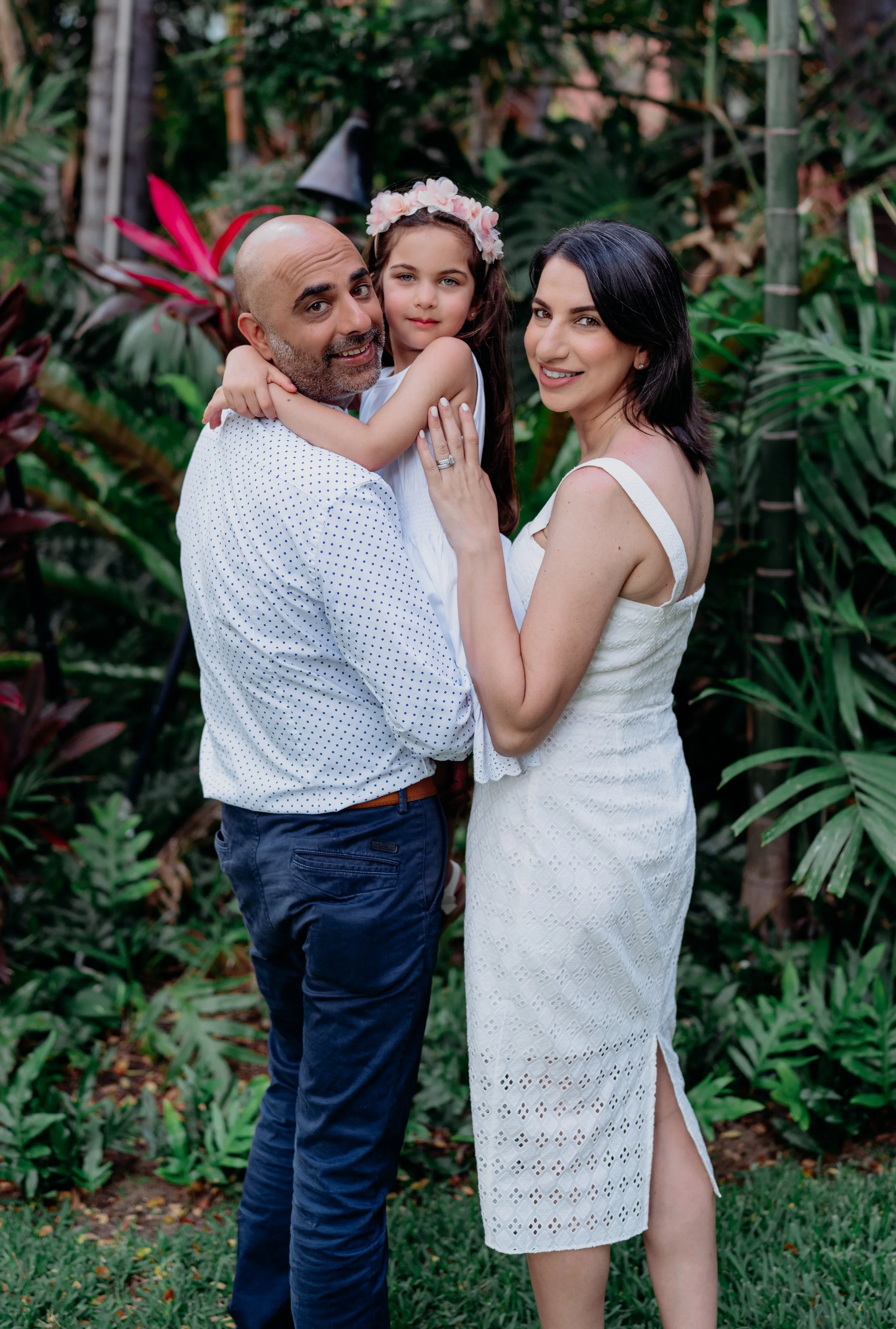 Family of three in a lush green garden, with the father holding his young daughter who is wearing a flower crown and smiling, while the mother stands close, holding the daughter's arm, all dressed in light-colored summer clothing.
