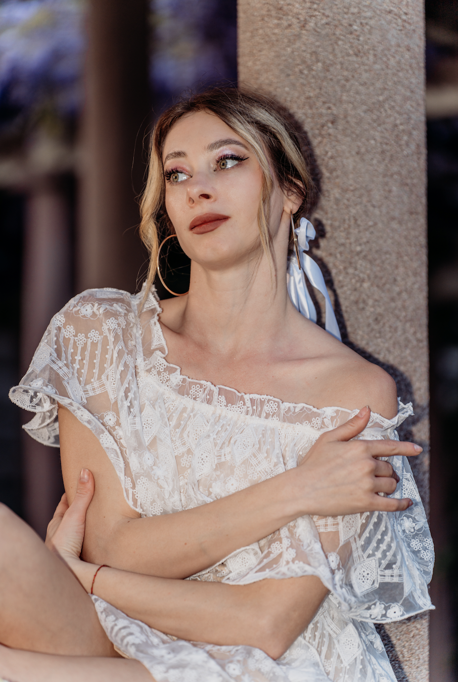 A woman with blonde hair, makeup, and hoop earrings, wearing a white lace off-shoulder dress, leaning against a stone column.