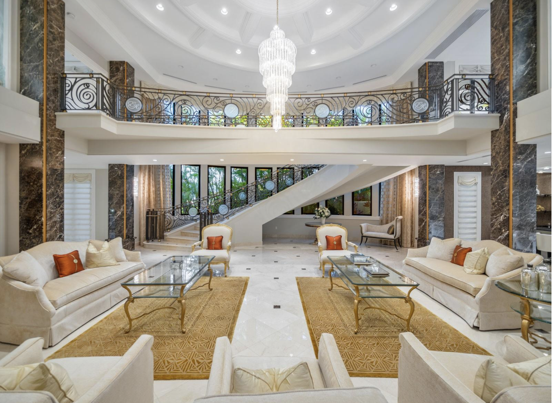 Luxurious hotel lobby with cream sofas, glass coffee tables, and elegant armchairs, featuring a grand staircase, a chandelier, and large windows with curtains.