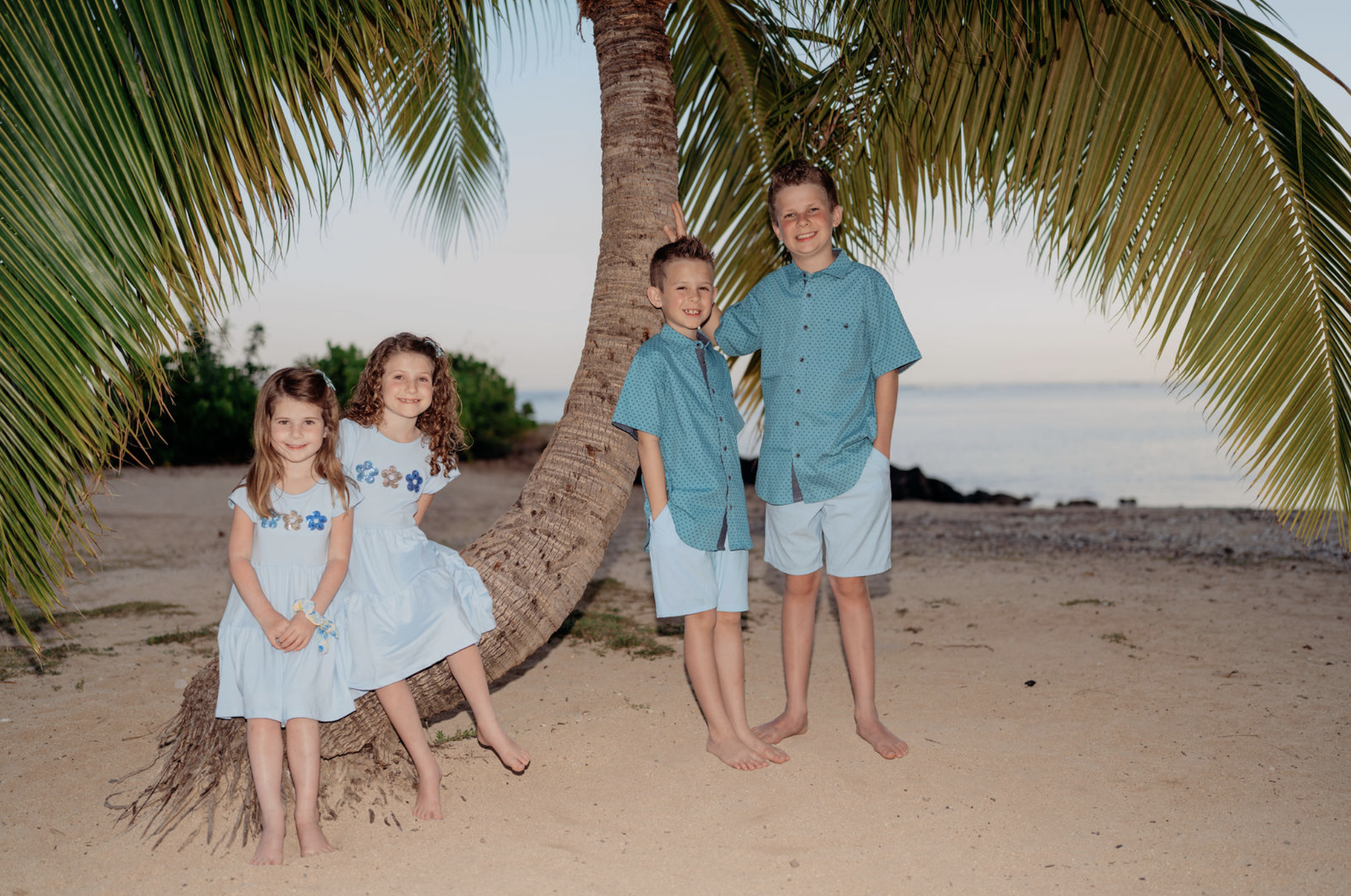 Four children smiling on a beach beside a leaning palm tree, with water in the background.