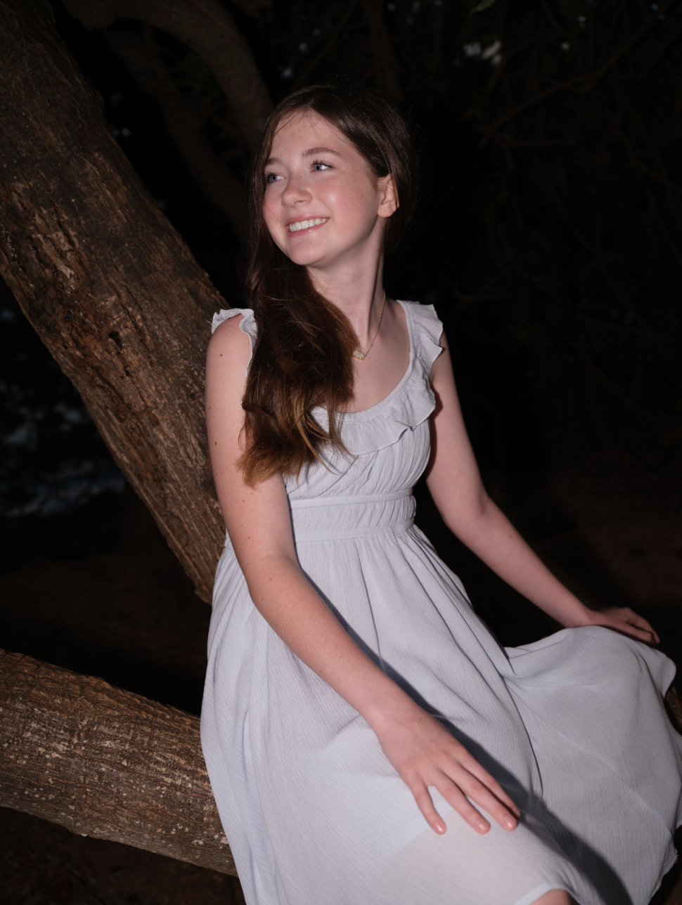A young woman with long brown hair sitting outdoors on a tree branch, wearing a white dress, smiling and looking off to the side.