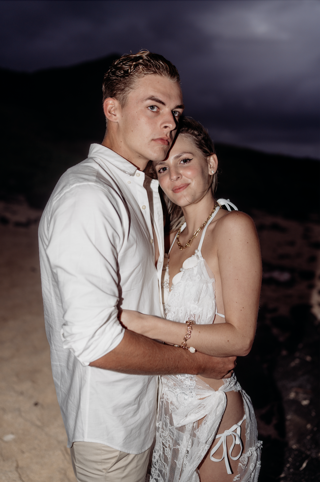 A couple embracing on a beach at dusk, both wearing white clothing.