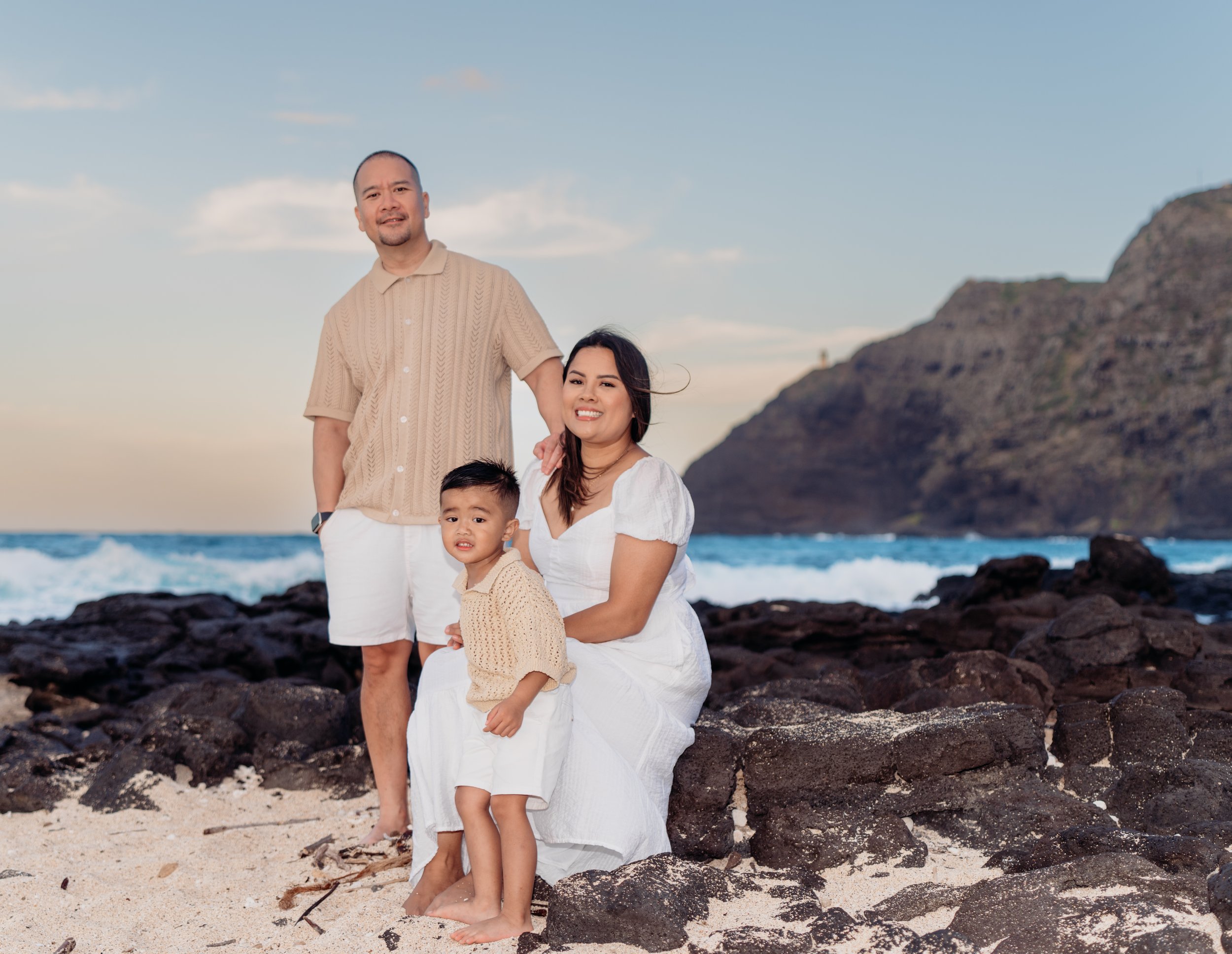 Family of three on a rocky beach with ocean and cliffs in the background, with a man standing and a woman and child sitting on rocks, all smiling and dressed in light-colored summer clothing.