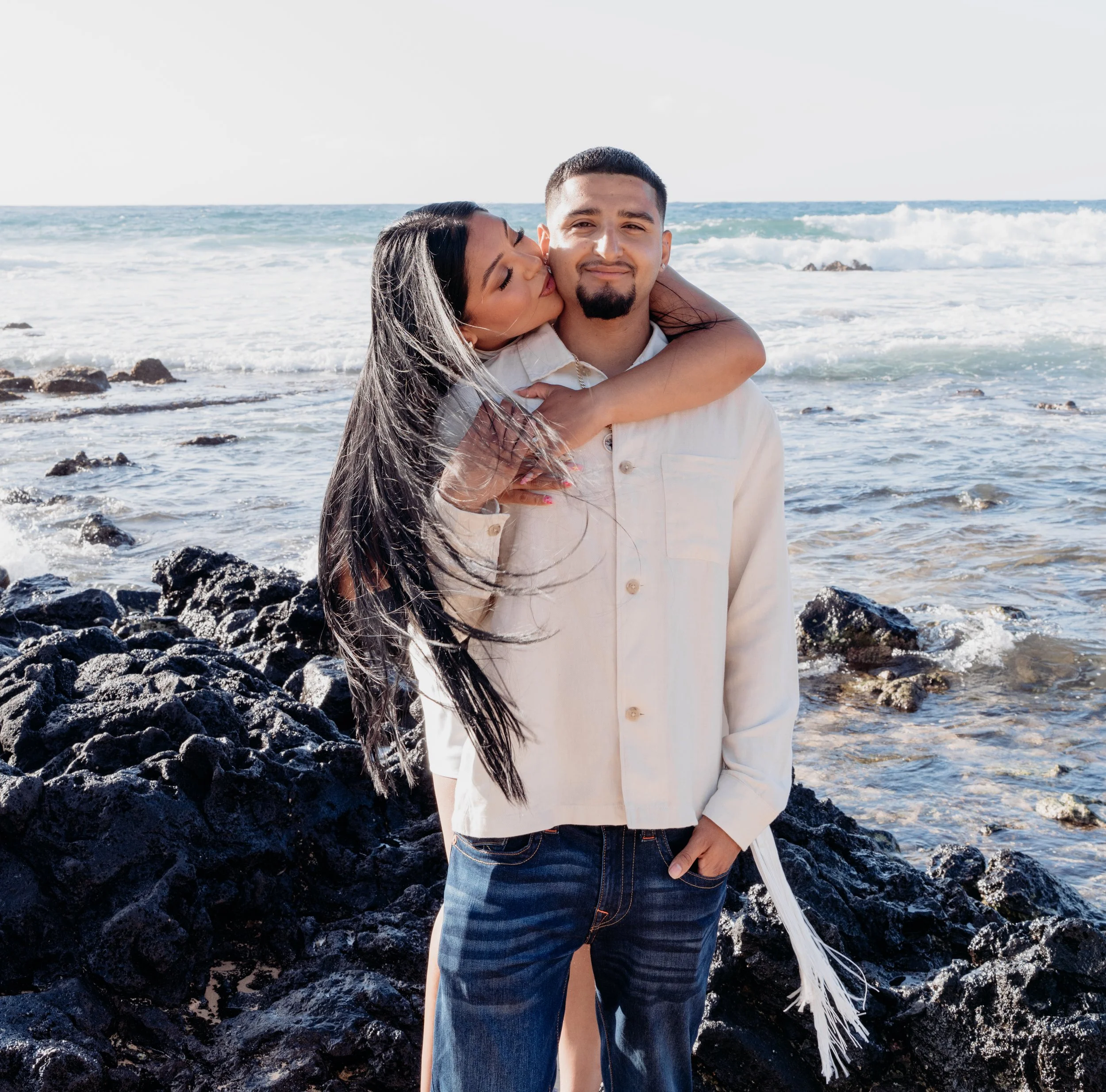 A woman kissing a man on the cheek while hugging him from behind on a rocky beach.