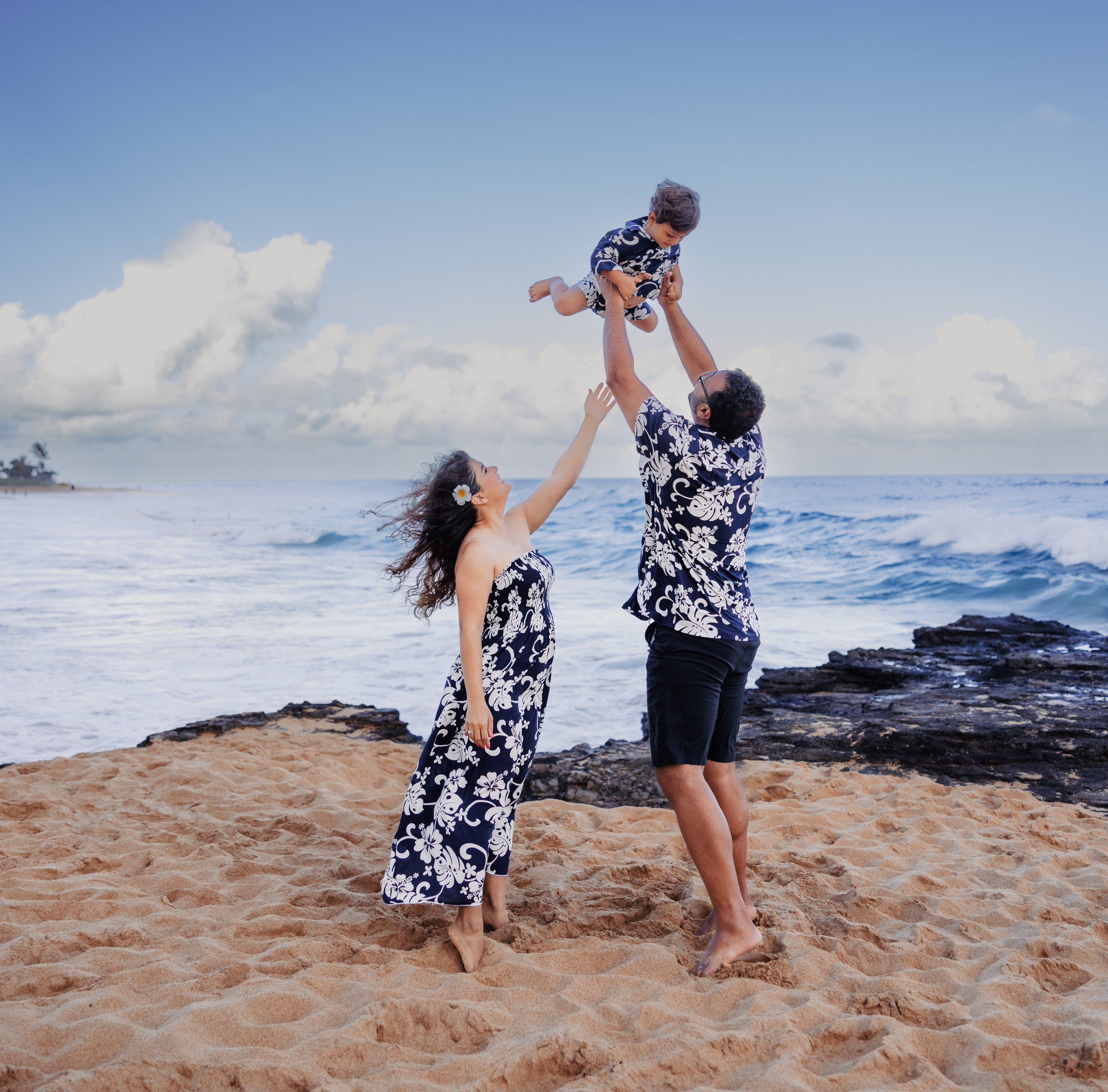 Family on the beach in matching Hawaiian shirts, playing and lifting a young boy in the air with ocean and cloudy sky in the background.