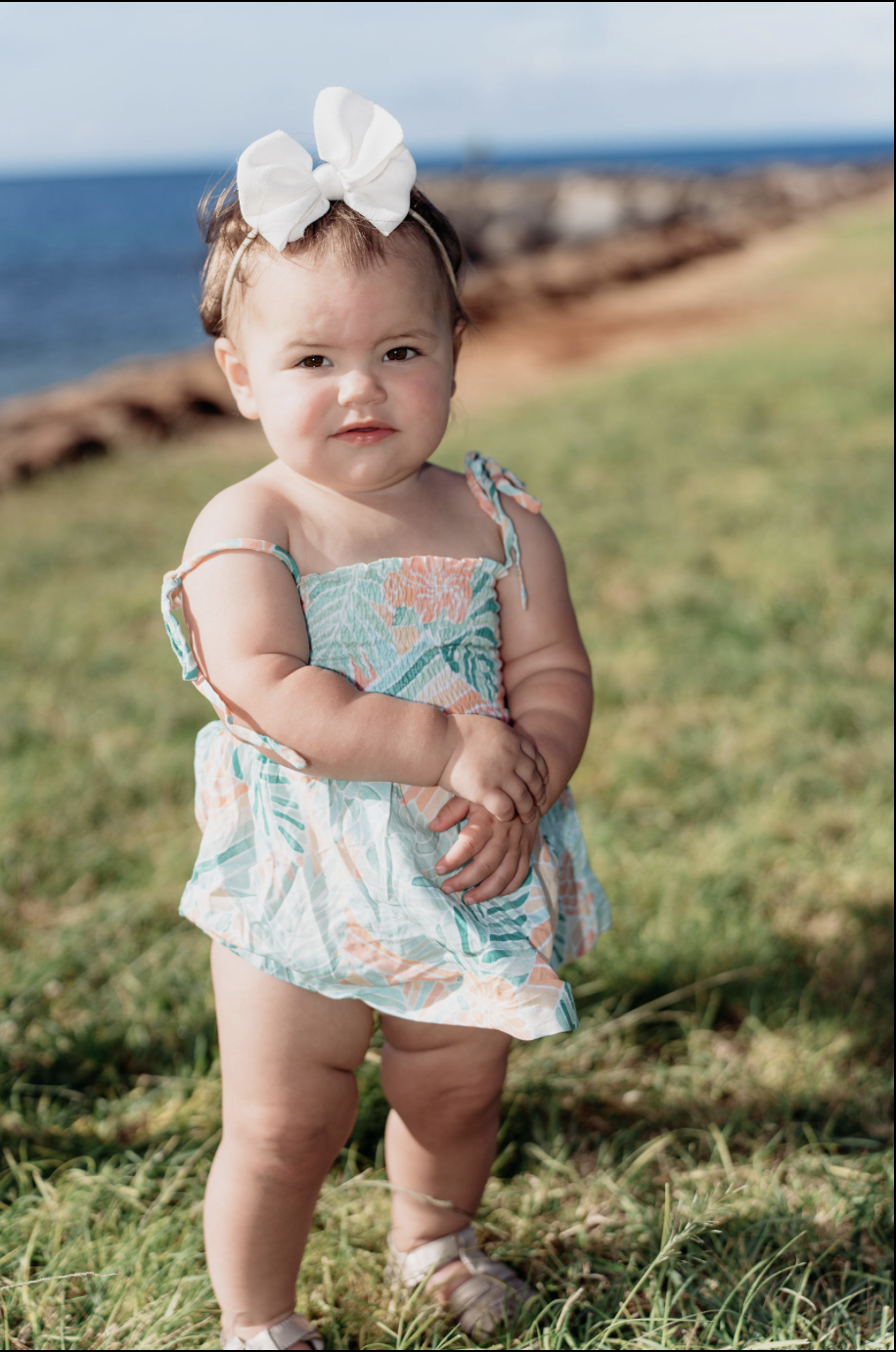 A young girl with a white bow in her hair, wearing a colorful floral dress, standing on grass near the ocean.