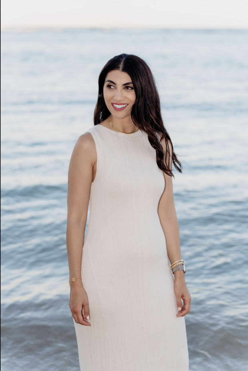 A woman with dark hair smiling at the camera, wearing a sleeveless white dress, standing in front of a body of water at the beach.