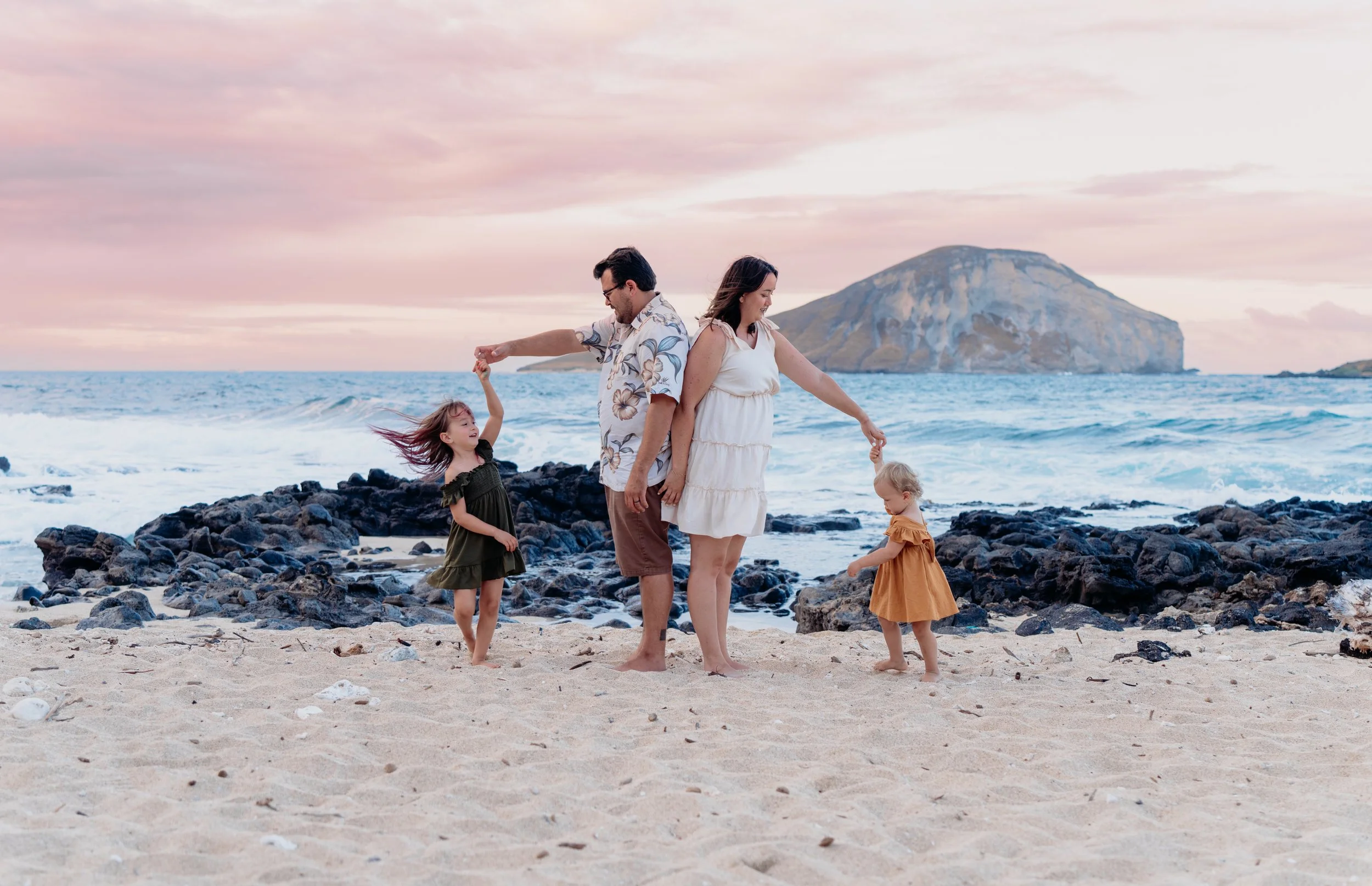 A family of four, two adults and two children, playing on a sandy beach with rocks, ocean waves, and a mountain in the background during sunset or sunrise.