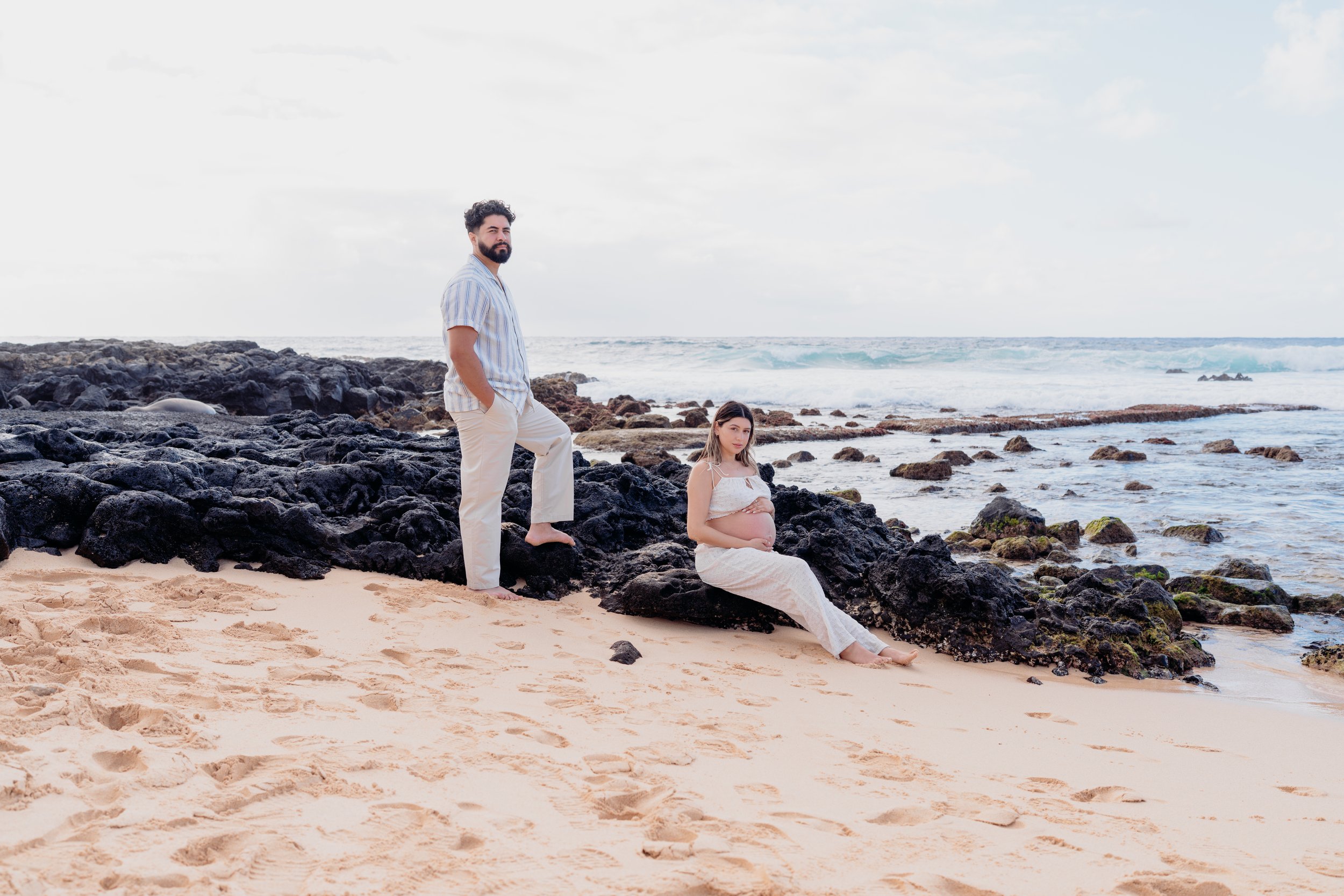 A pregnant woman in a white dress sitting on rocks by the beach with her partner standing nearby, overlooking the ocean.