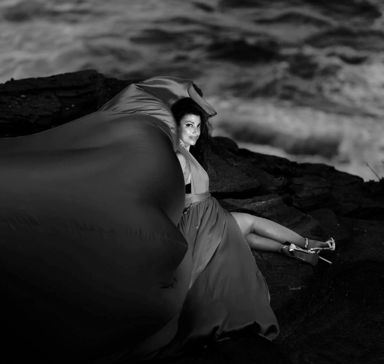 A woman sitting on a rocky surface near the water, wearing a dress and high heels, with a large umbrella draped over her shoulders.