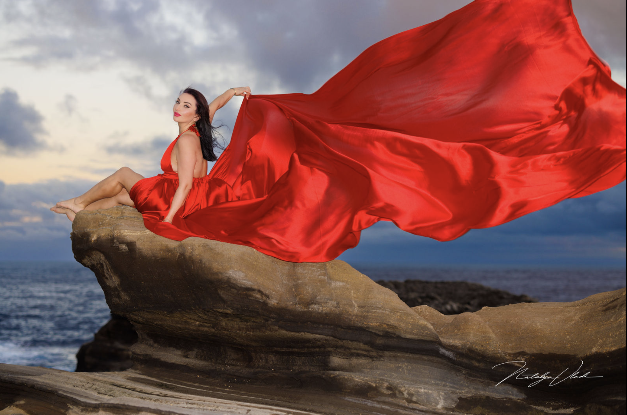 A woman in a flowing red dress sitting on a rock by the ocean, waving a long piece of red fabric in the wind during sunset.