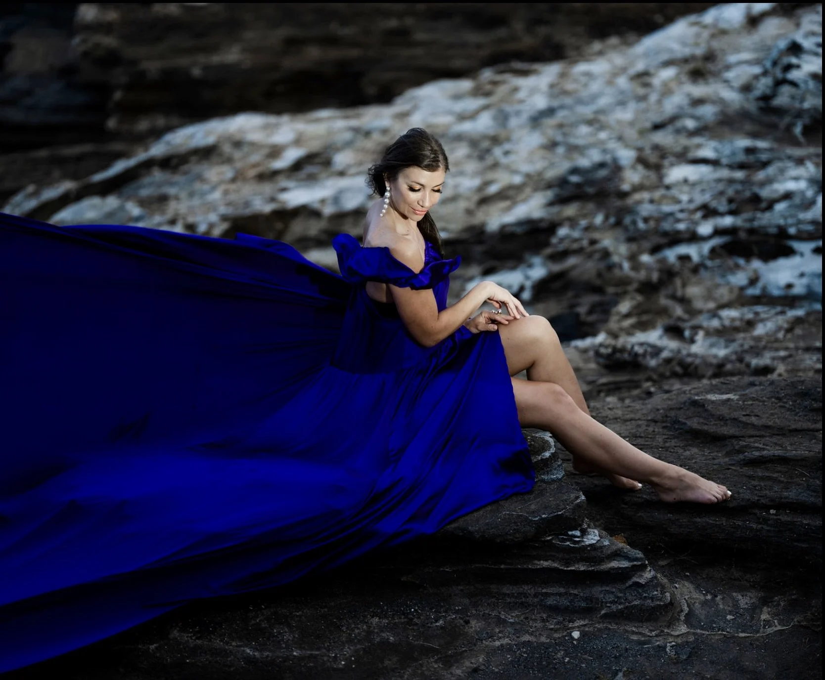 Woman sitting on a rocky surface wearing a flowing deep blue gown, with a natural landscape background.