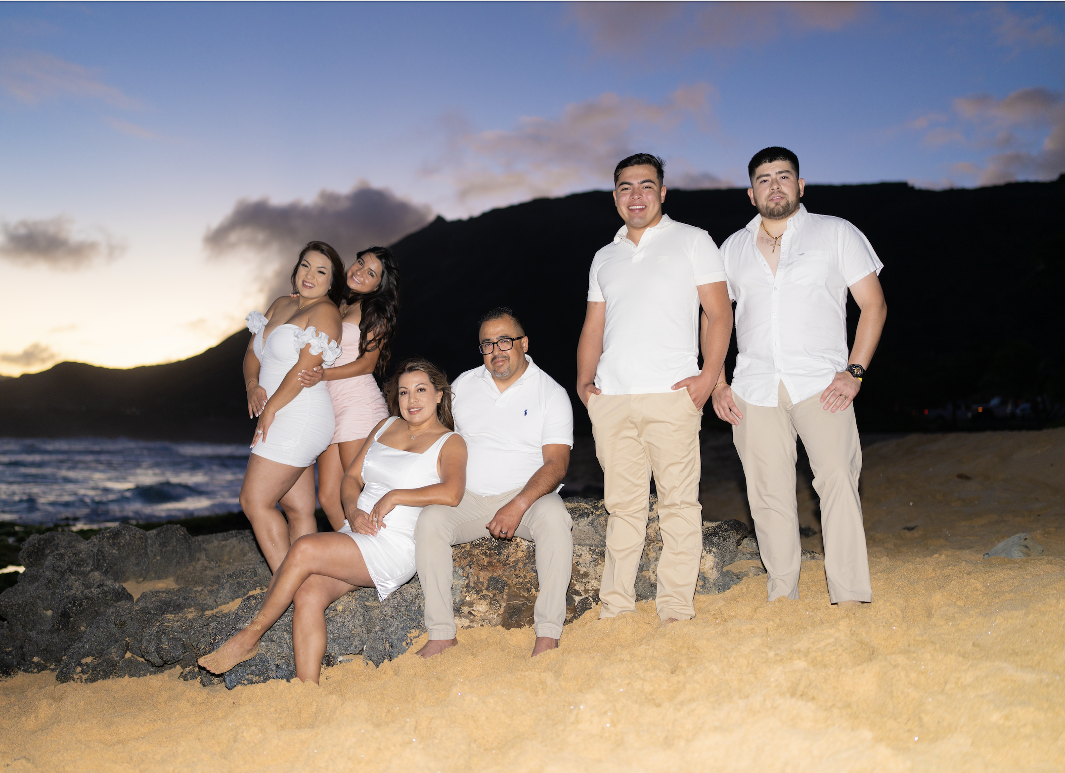 Group of six people on a beach during sunset, with a mountain and cloudy sky in the background. Four people are standing and two are sitting on rocks, all dressed in light-colored casual clothes.