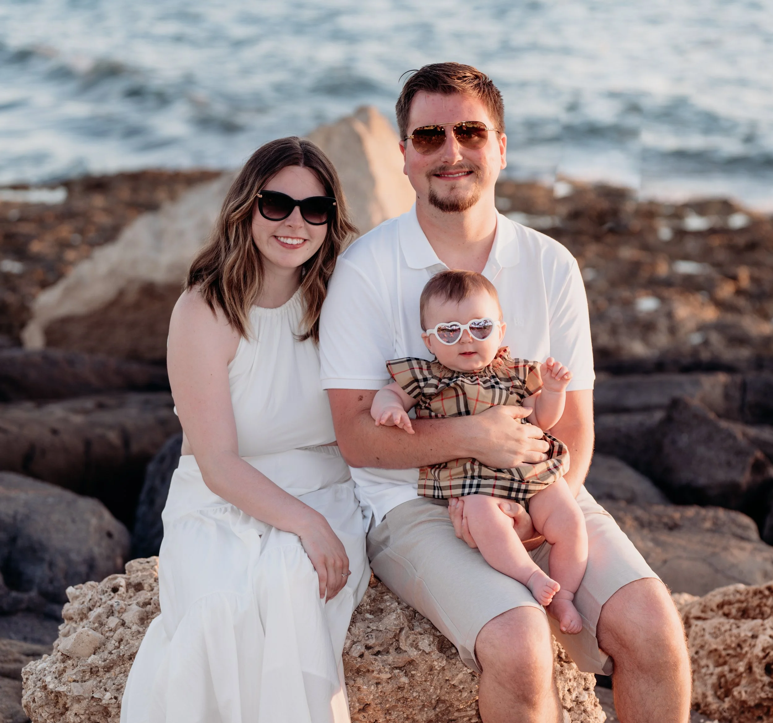 A family of three, a man, a woman, and a baby, sitting on rocks at the beach with the ocean in the background. They are all wearing sunglasses and smiling.