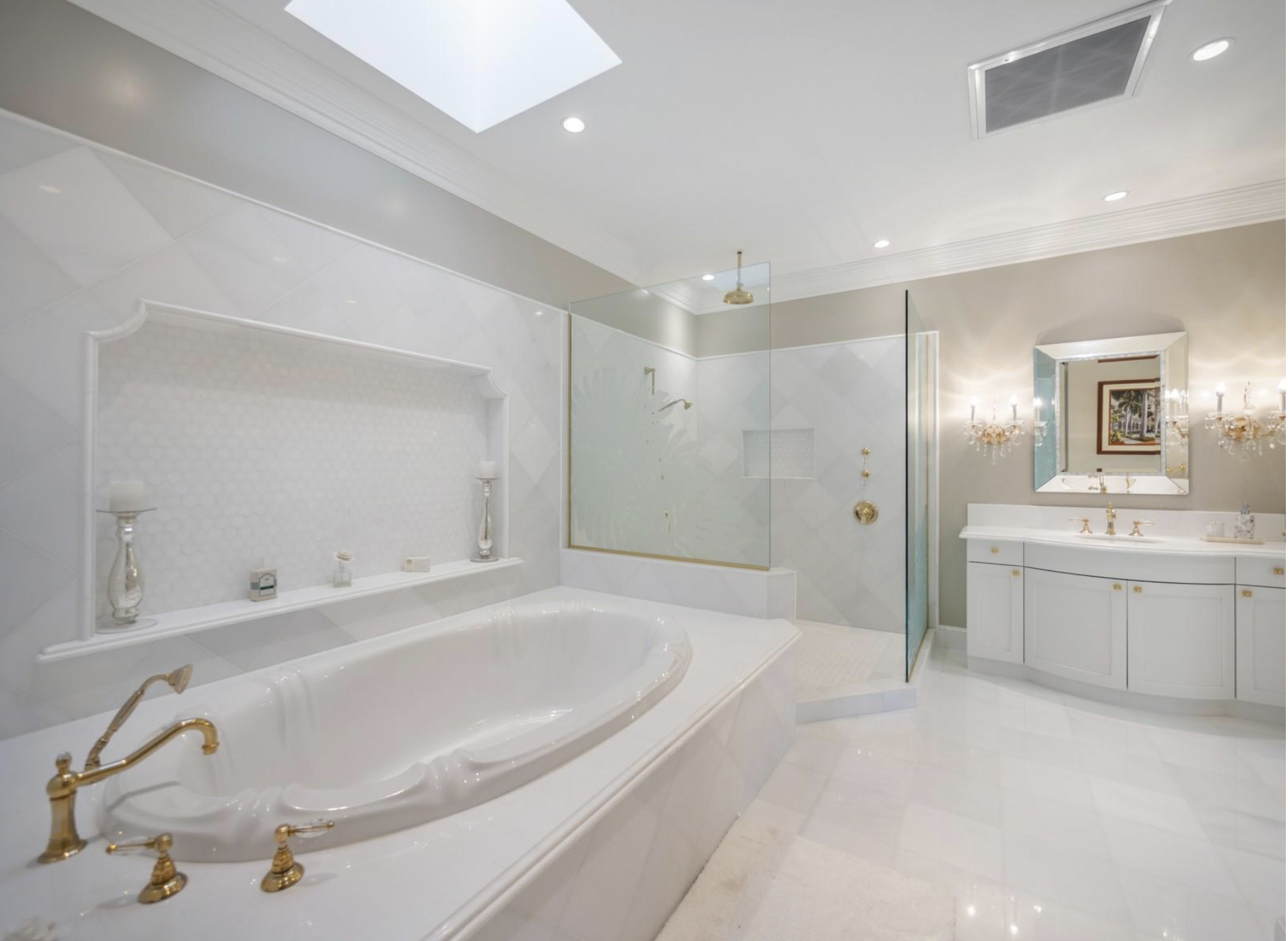 Elegant bathroom featuring a large bathtub with gold fixtures, a spacious glass shower with gold accents, a white vanity with a large mirror, and chandelier lighting.