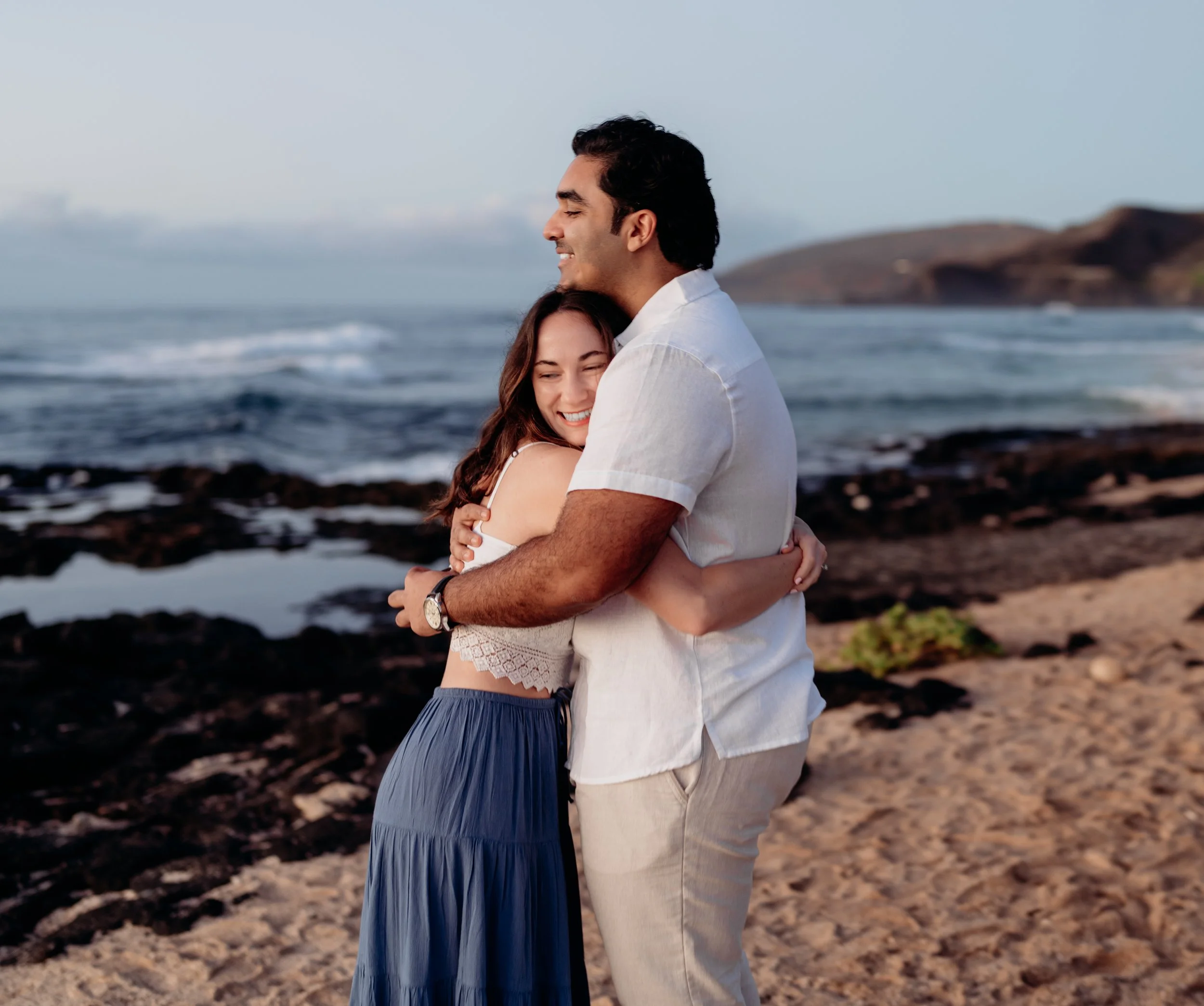 A man and woman hugging on a rocky beach with ocean waves in the background, smiling and enjoying each other's company.