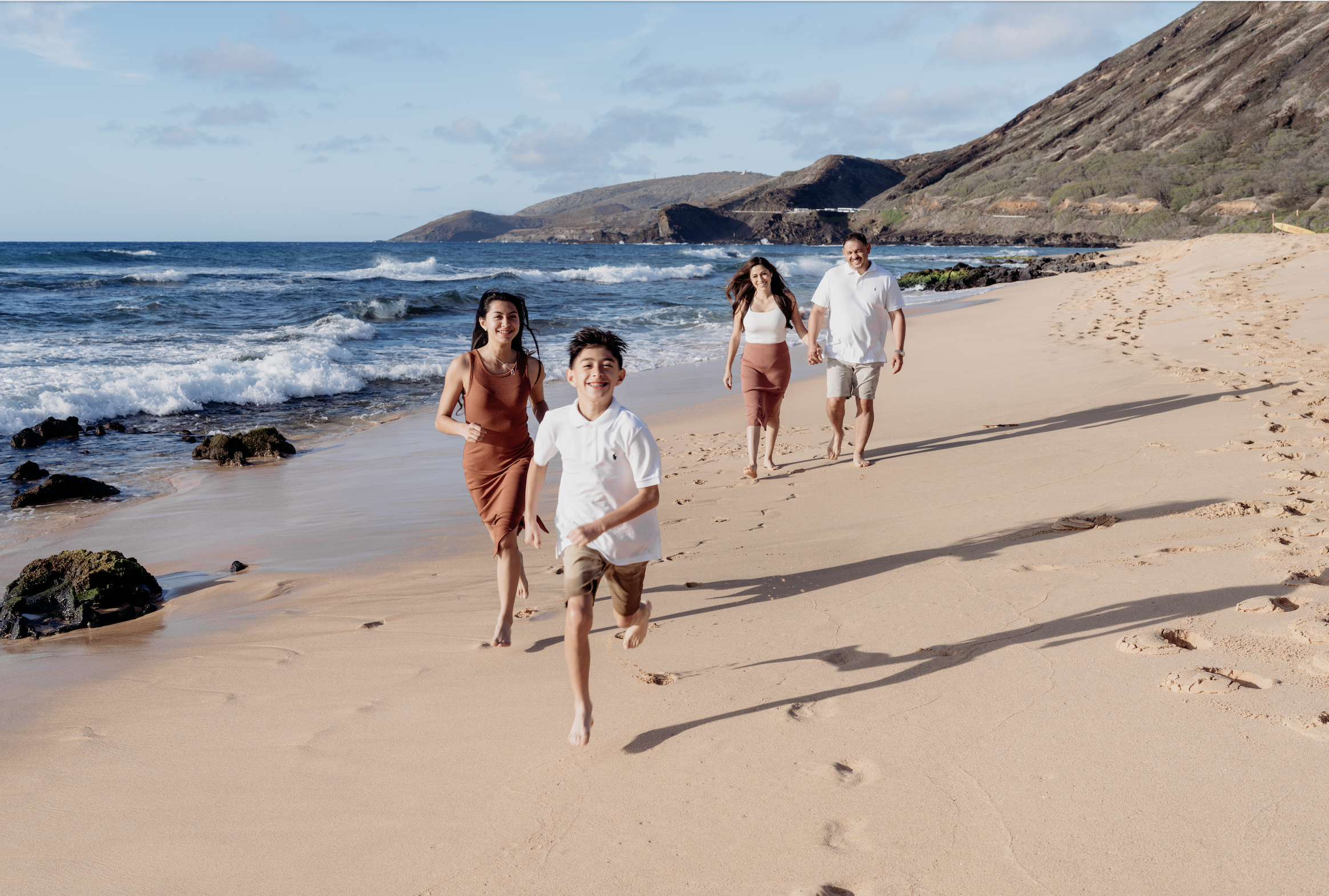 A family of four runs barefoot on the sandy beach near the ocean, with waves crashing and hills in the background on a sunny day.