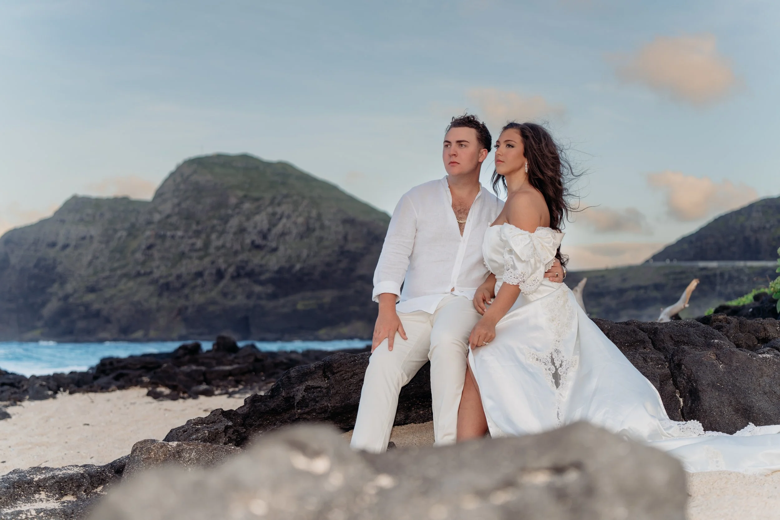 A couple dressed in white clothing sitting on rocks on a beach with a mountainous landscape in the background.