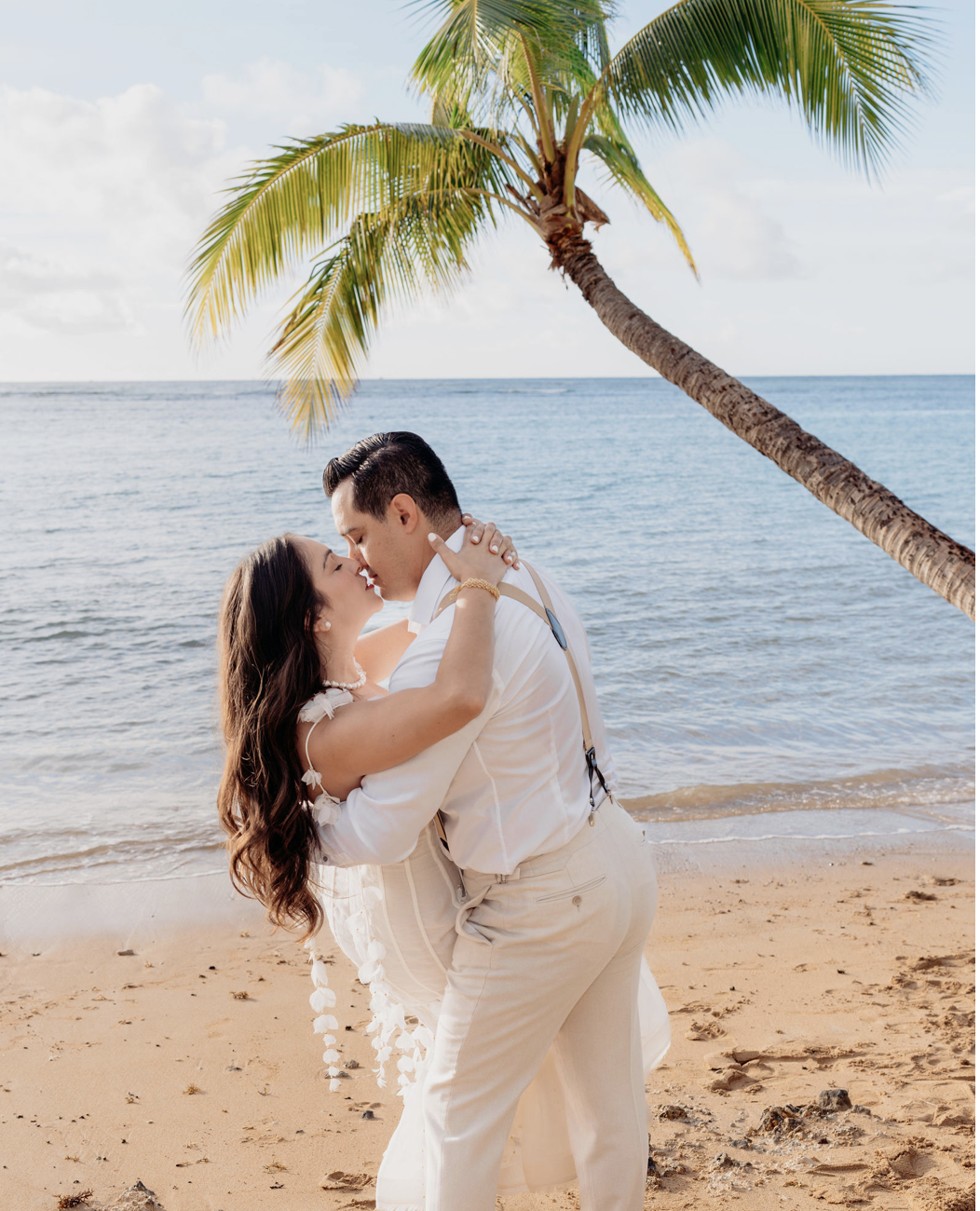 A couple kissing on a beach with a palm tree in the background, wearing white wedding attire, ocean water, sandy shore, and partly cloudy sky.