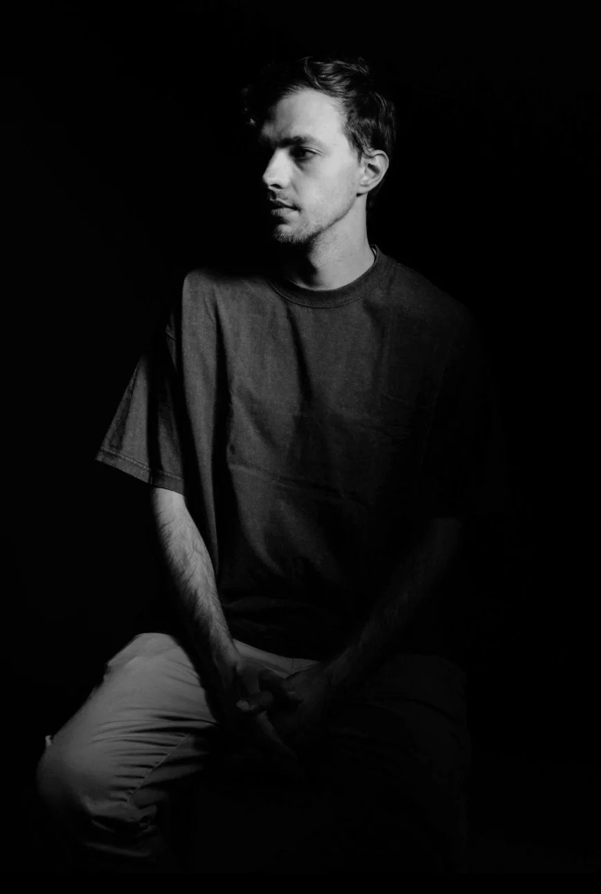 Black and white photo of a young man sitting against a dark background, looking to the side with a serious expression.