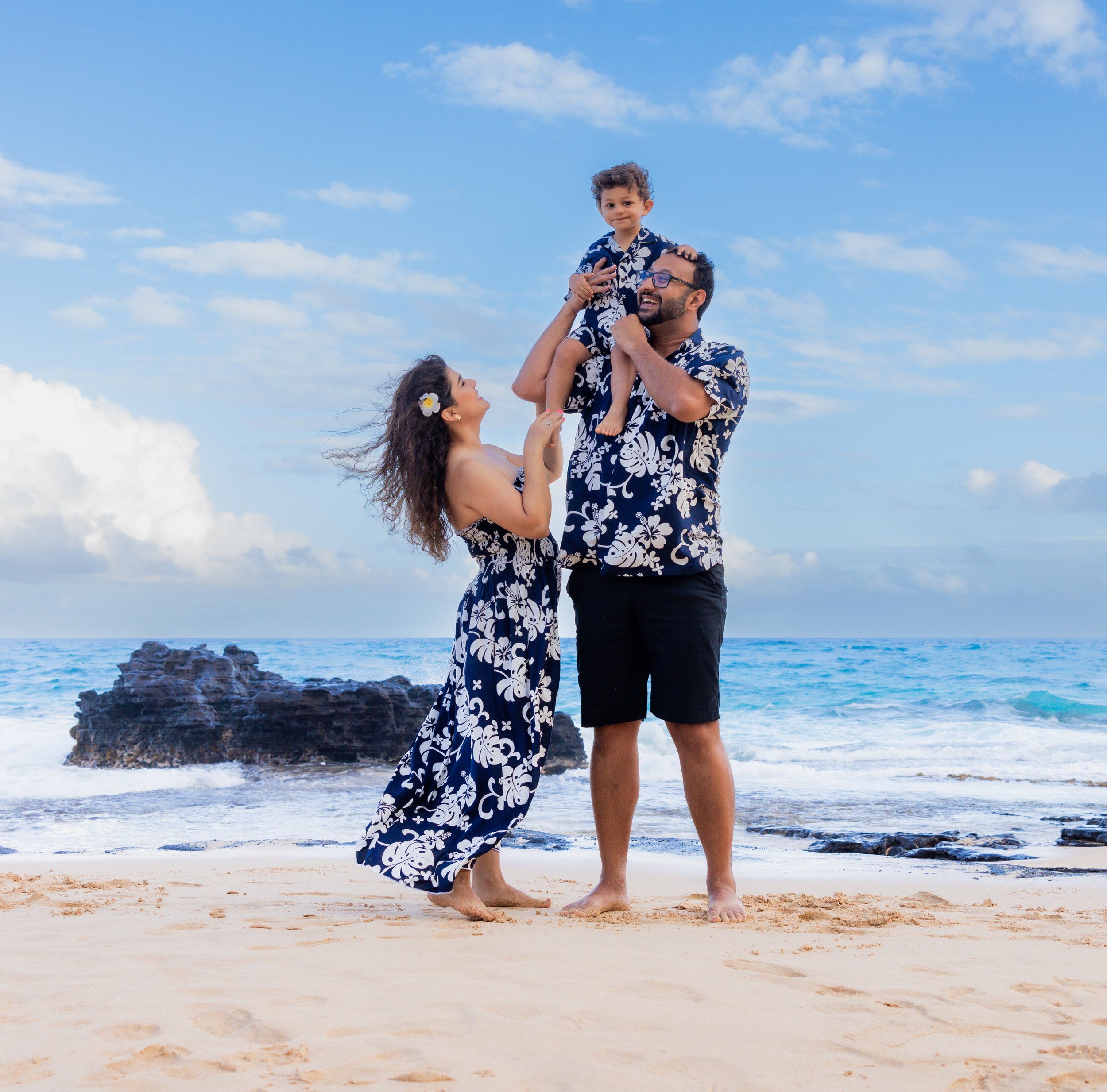 Family of three on the beach, wearing matching Hawaiian shirts, with the ocean in the background. The father is holding a young boy on his shoulders, the mother is touching the boy's hand, and all are smiling.