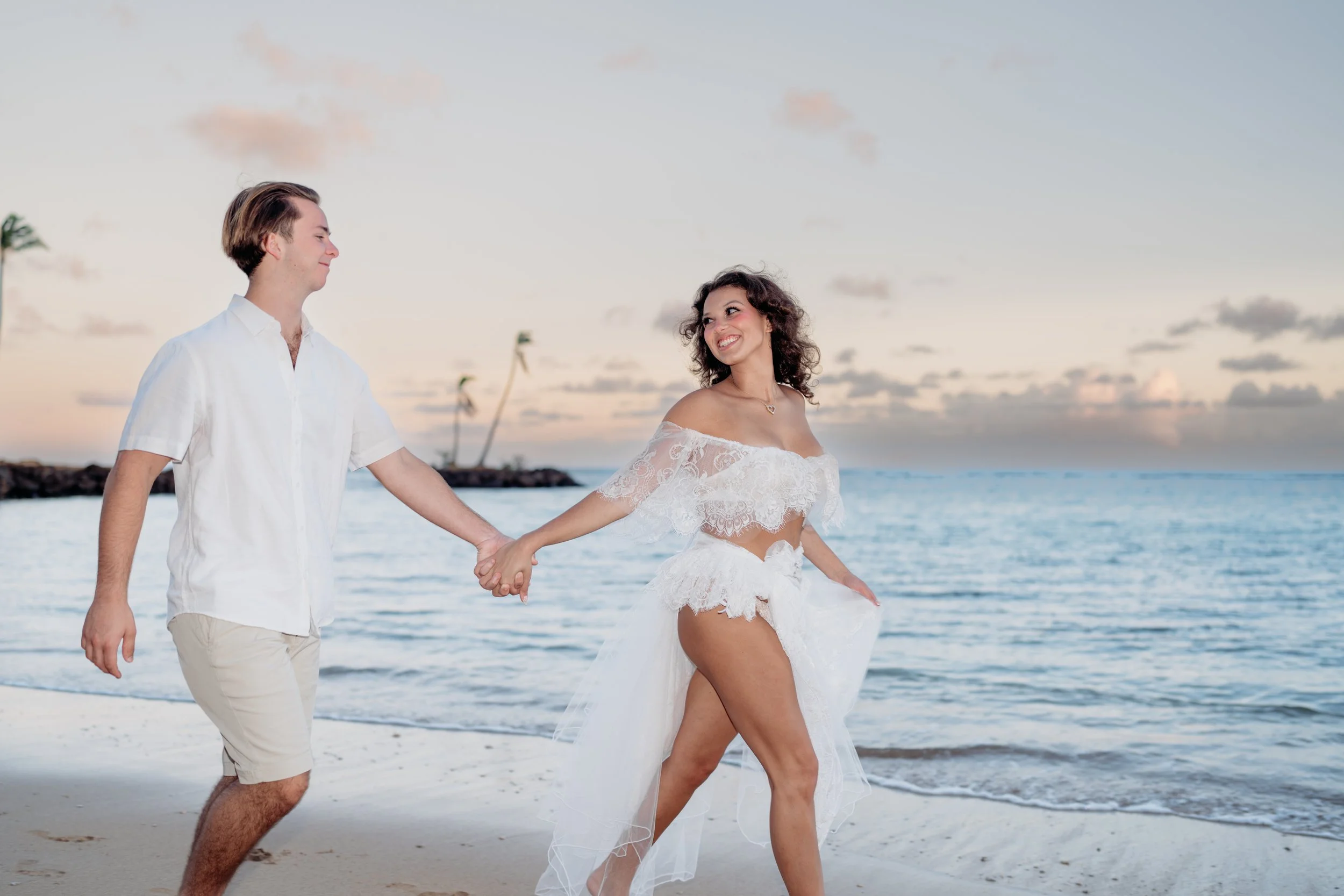 A couple holding hands on the beach at sunset, with palm trees and ocean in the background.