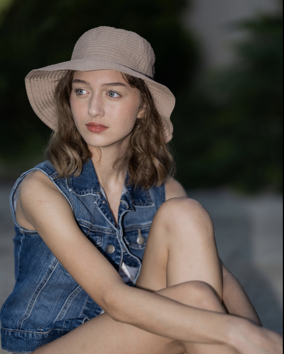 Young woman with curly brown hair wearing a large beige sunhat and denim sleeveless jacket, sitting outdoors with a blurred green background.