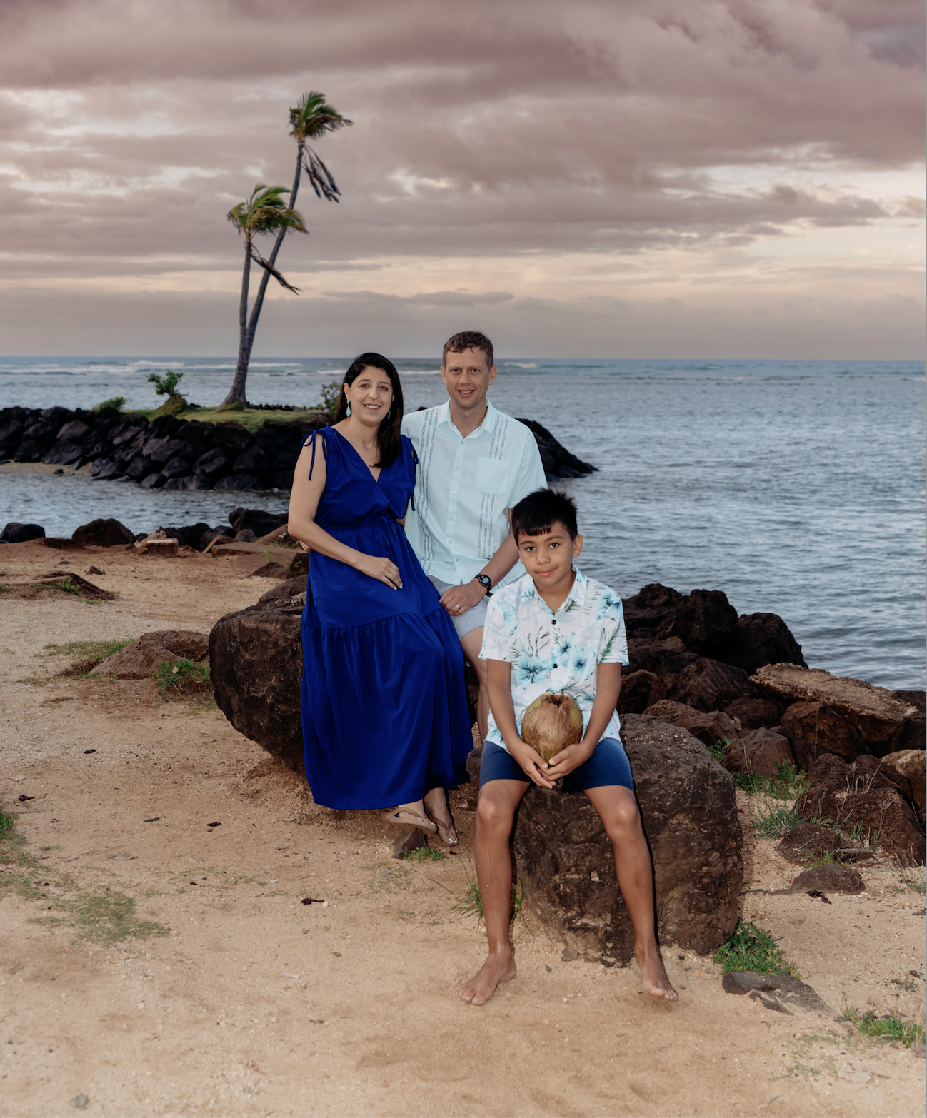 A family of three posing on a beach in front of the ocean and a small island with two leaning palm trees at sunset.