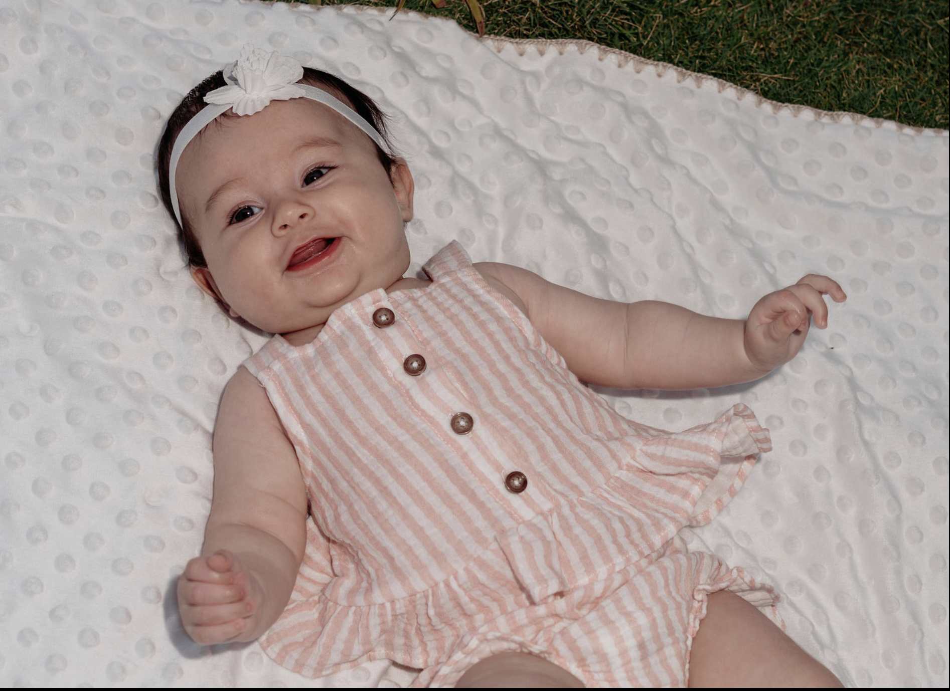 An adorable baby girl lying on a white dimpled blanket outdoors, wearing a light pink striped dress with brown buttons and a white headband with a bow. She is smiling and looking happy.