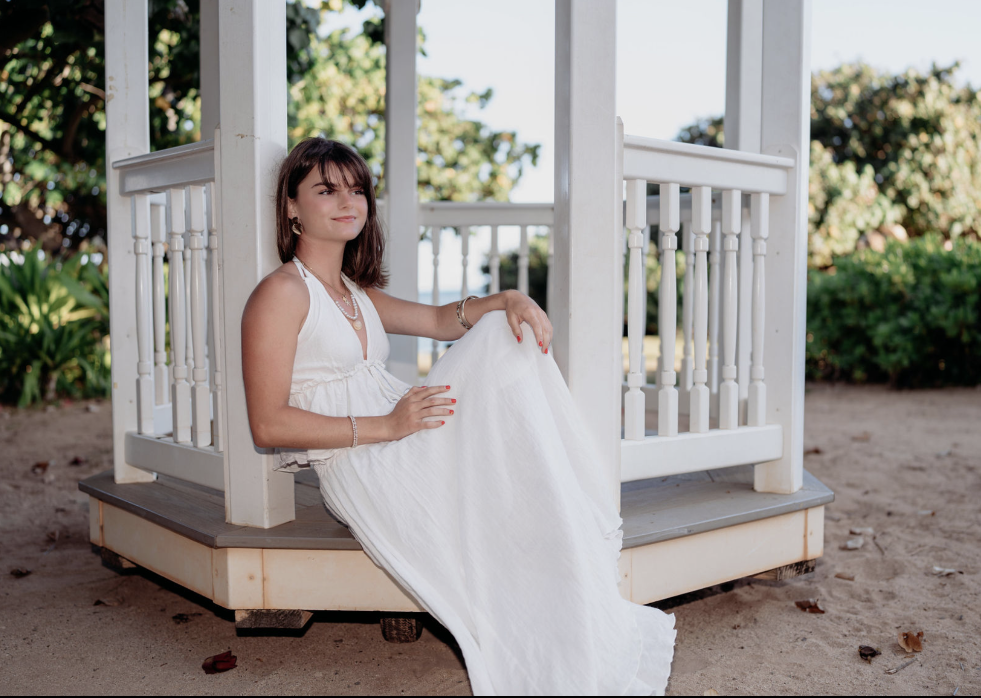 A young woman in a white dress sitting on a small wooden gazebo on a sandy beach with green bushes and trees in the background.