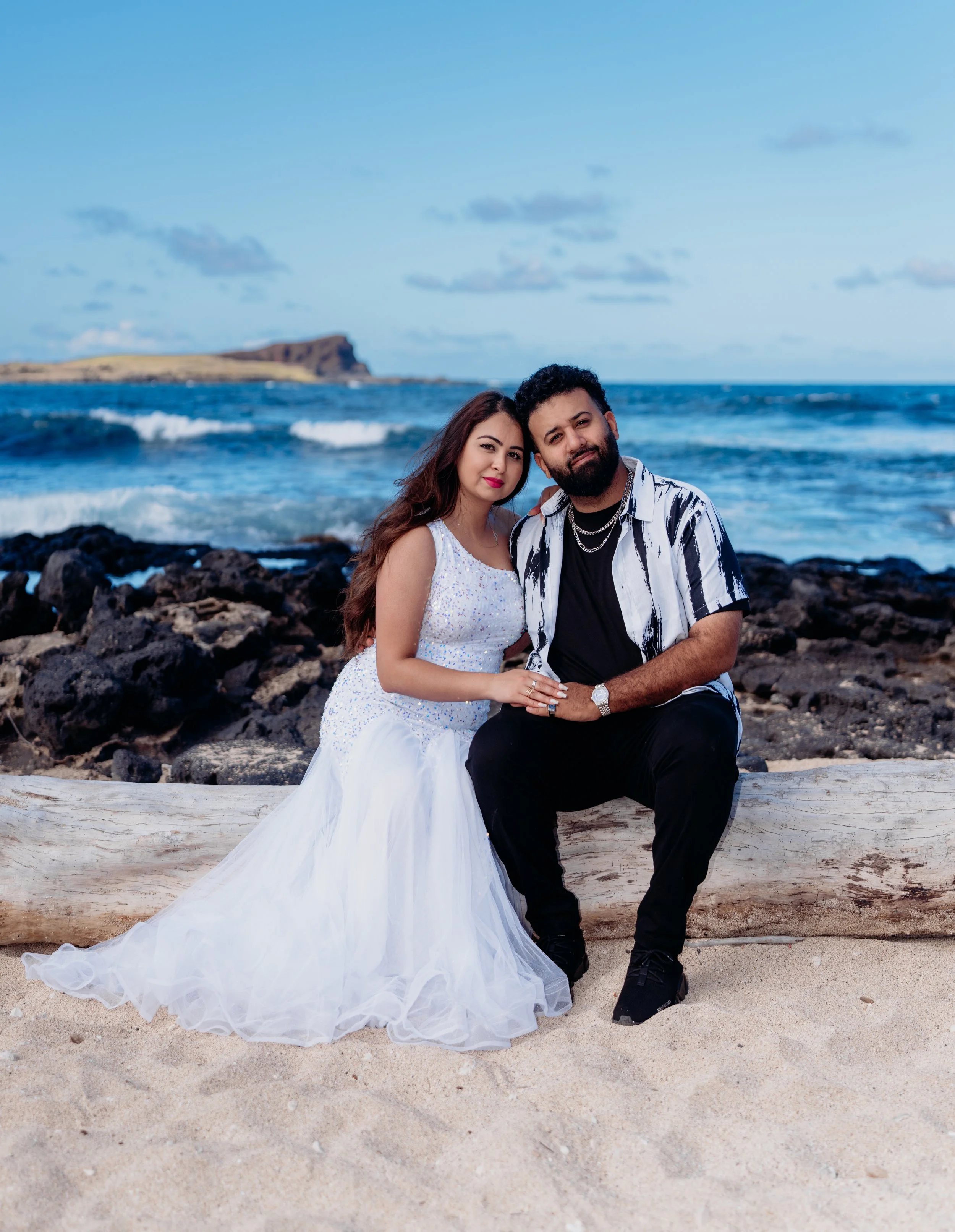 A couple sitting on a log at the beach, with the ocean and rocky shoreline in the background, during daytime.