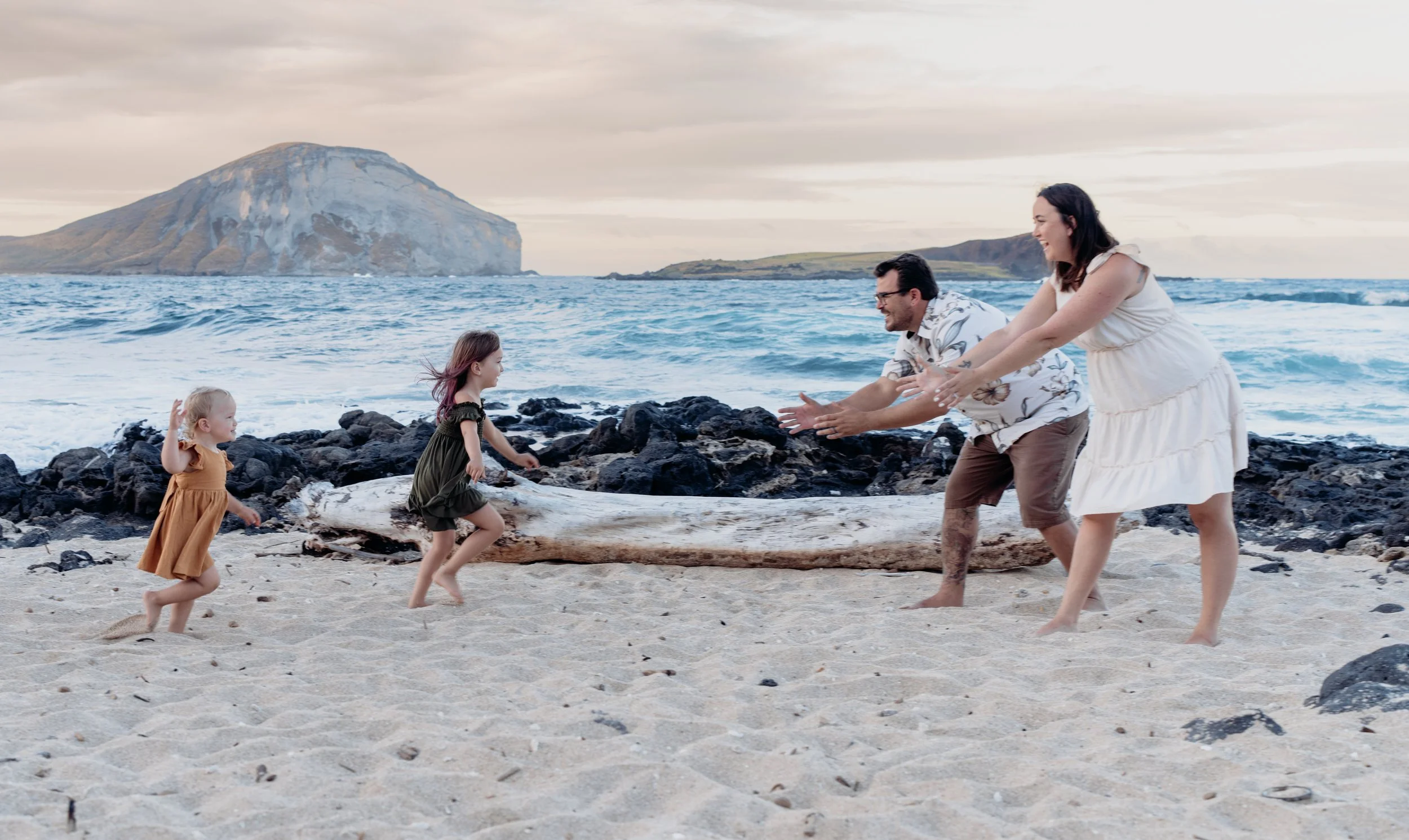 A family of four playing on a beach with rocks and driftwood, with water and a mountain in the background.