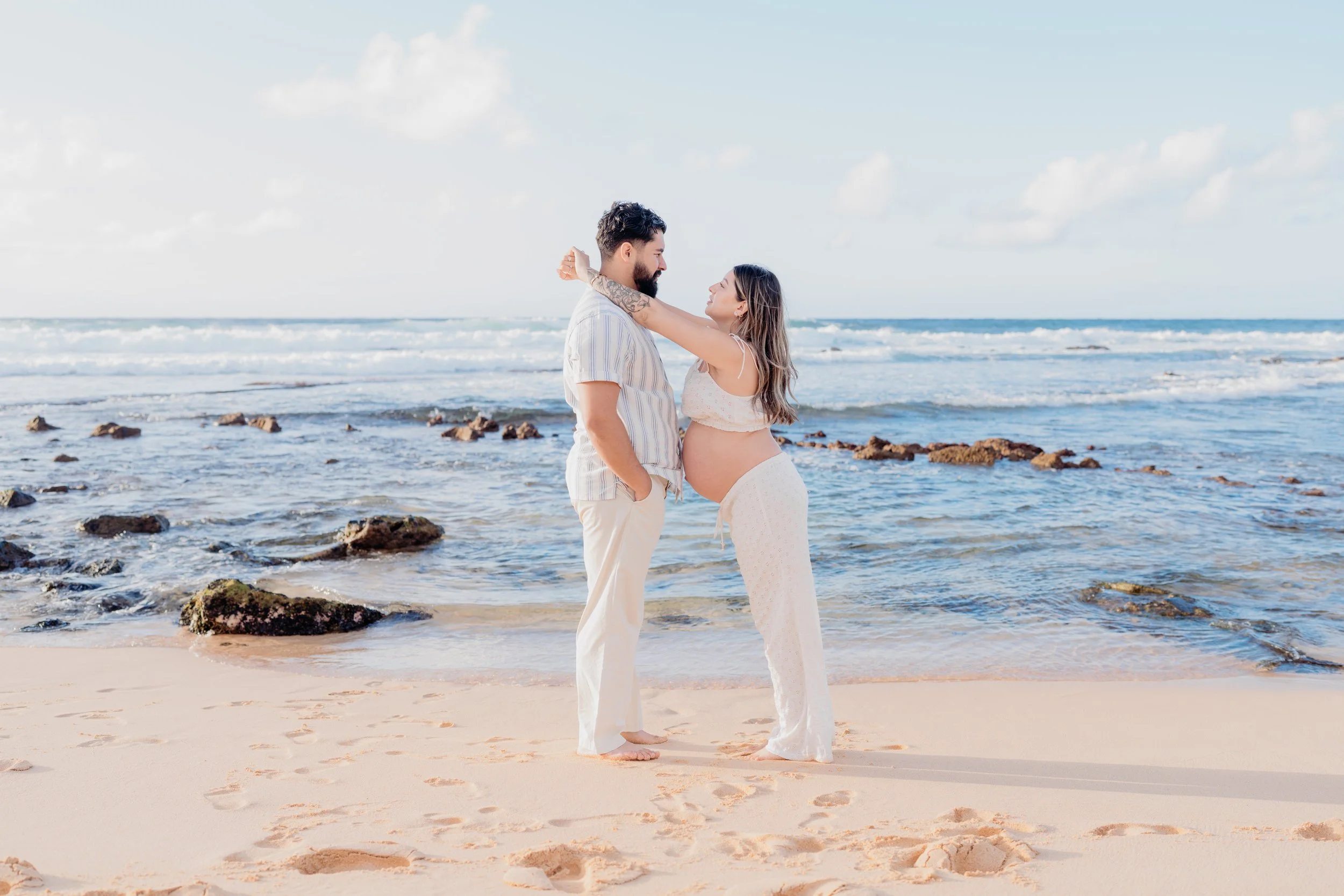 A pregnant woman and a man standing on a beach, looking at each other lovingly, with the ocean and sky in the background.