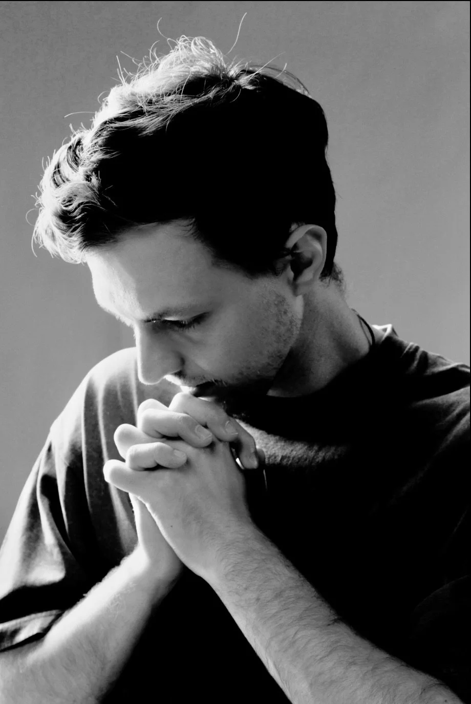A black and white photo of a young man with dark hair praying with hands clasped together and head bowed.