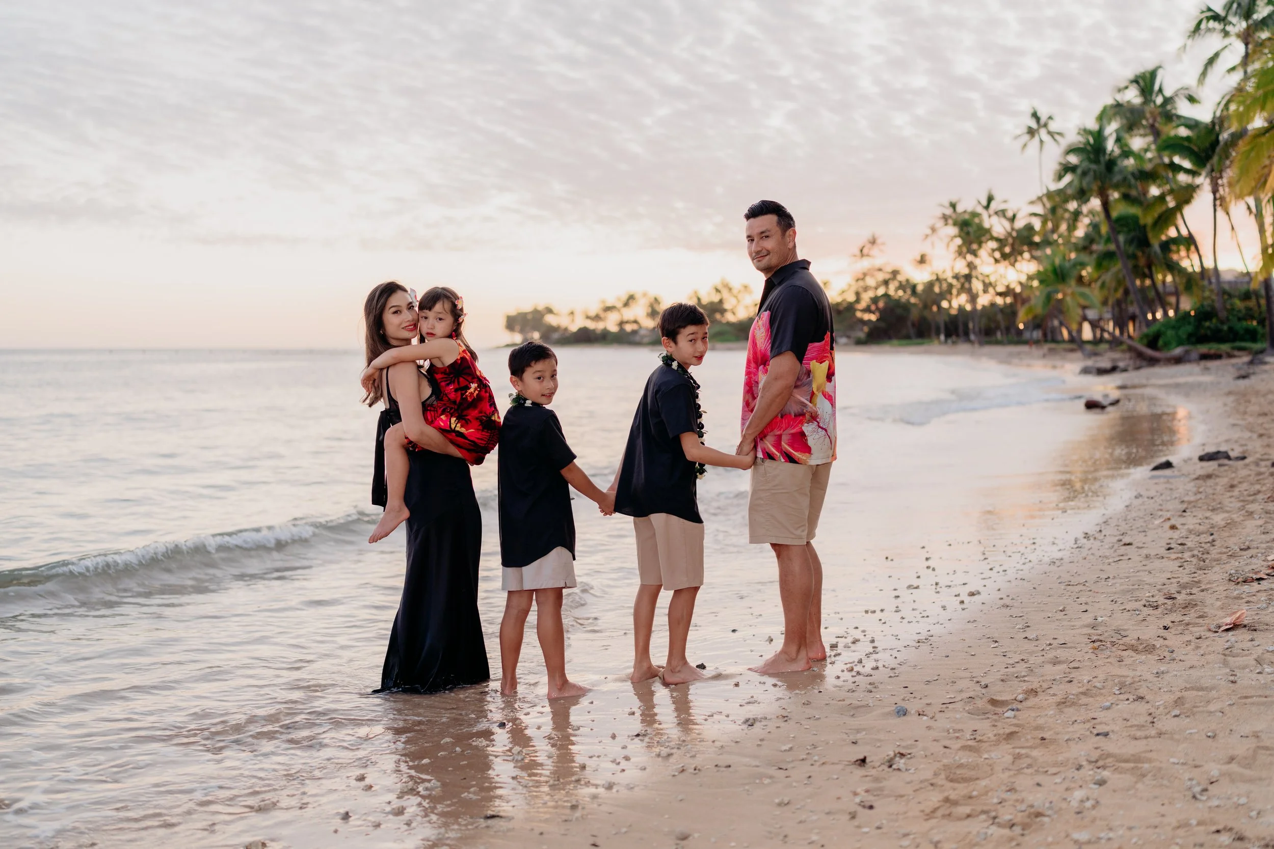 A family of six standing on a beach holding hands during sunset, with palm trees and the ocean in the background.