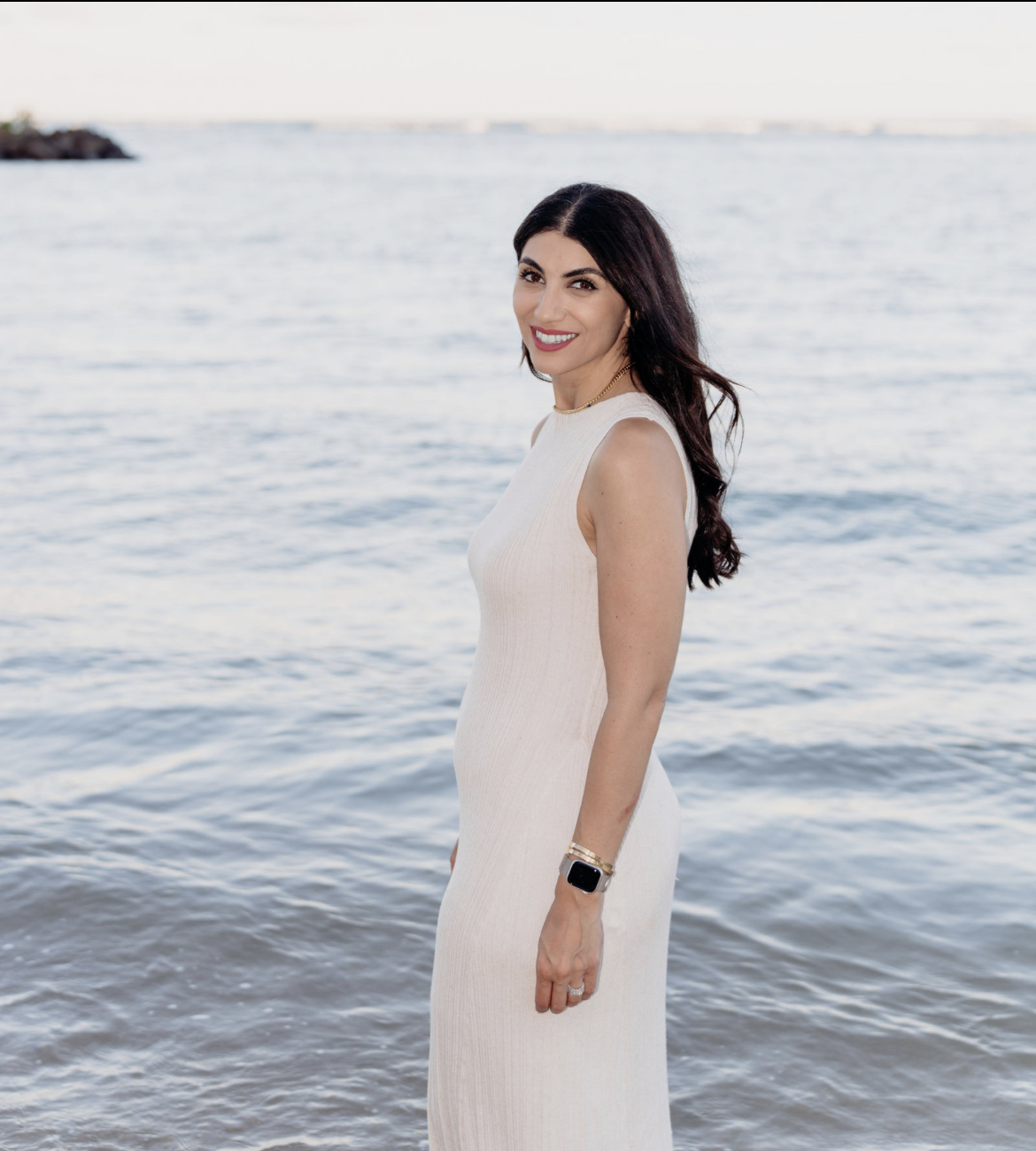 A woman in a white sleeveless dress smiling by the water with a distant shoreline in the background.