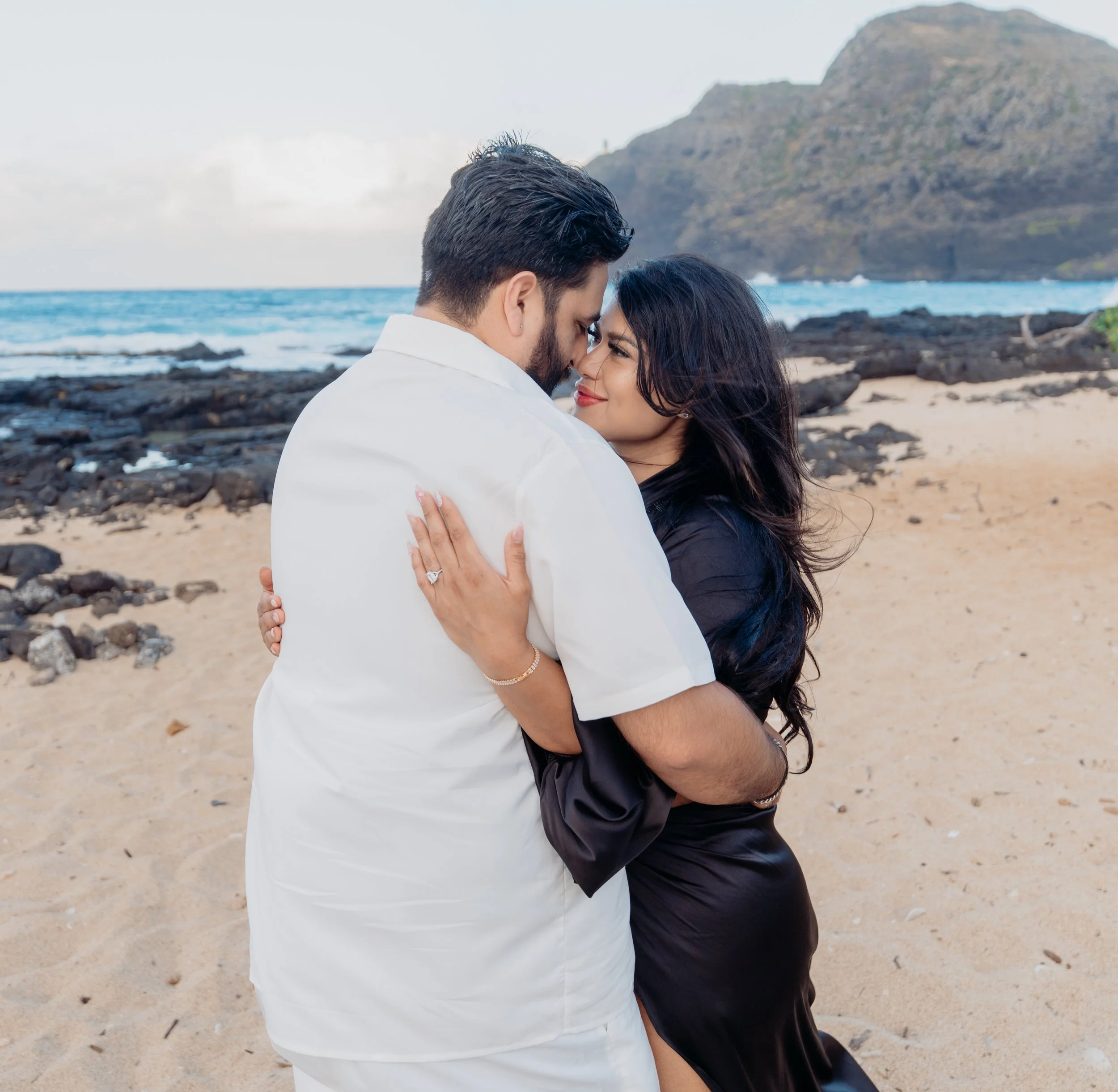 A couple embracing on a sandy beach with a rocky shoreline and a mountain in the background.