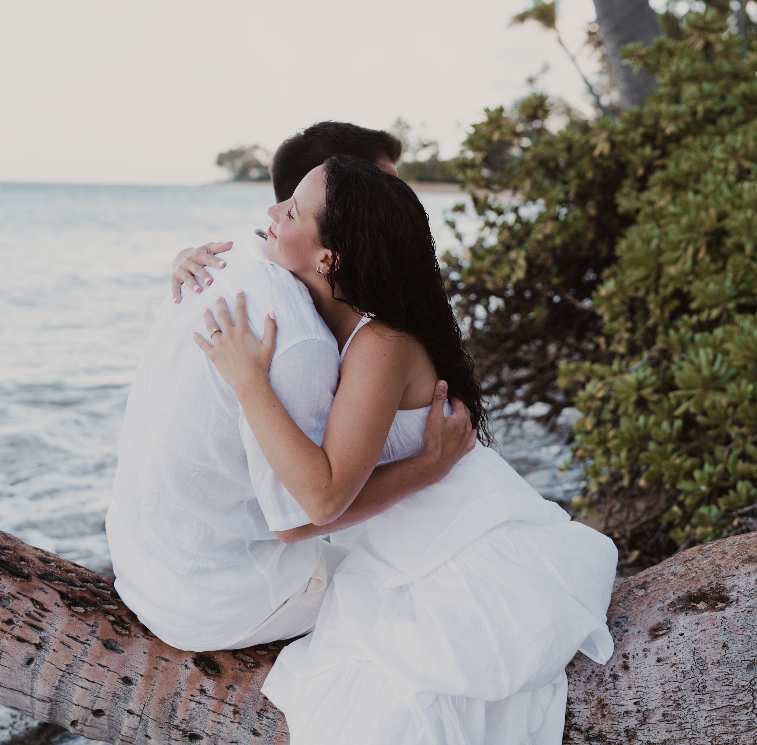 A couple hugging on a beach, sitting on a fallen tree trunk near the water, with greenery and trees in the background.