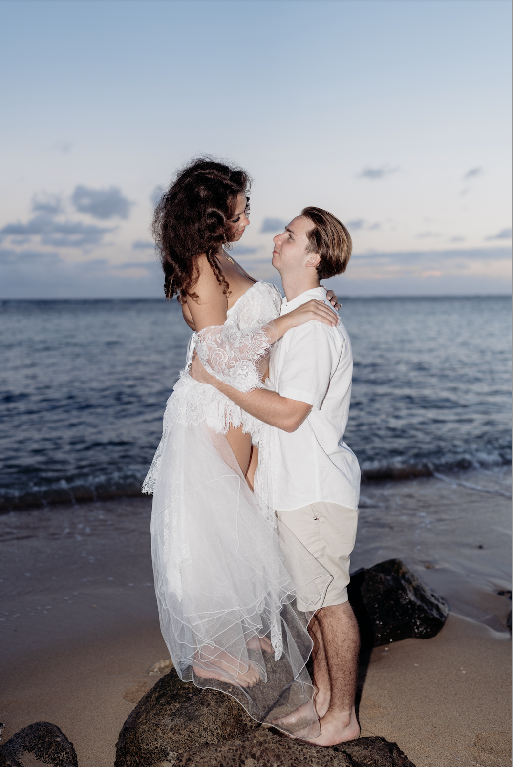 A couple in white clothing embracing on a rocky beach at sunset, with the ocean in the background.