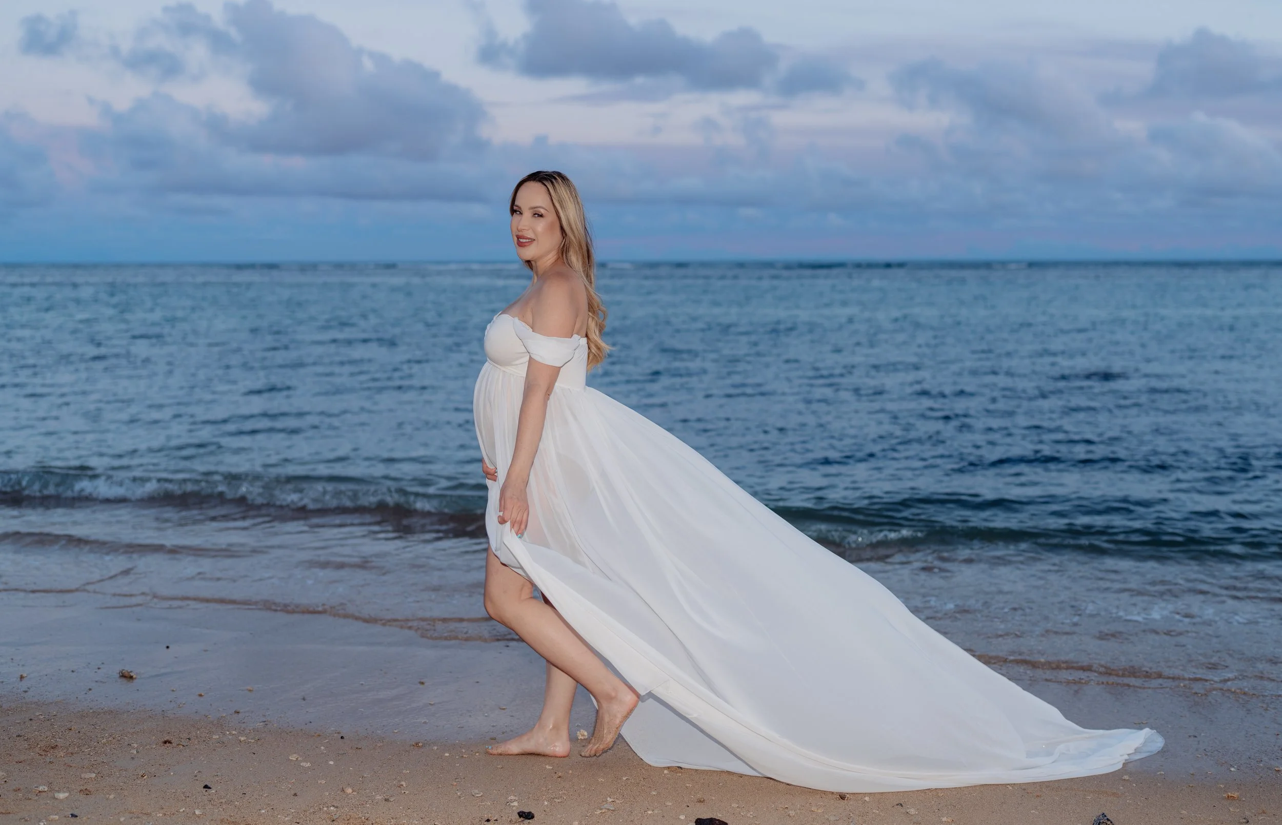 A pregnant woman in a white dress stands on a beach with the ocean and cloudy sky in the background.