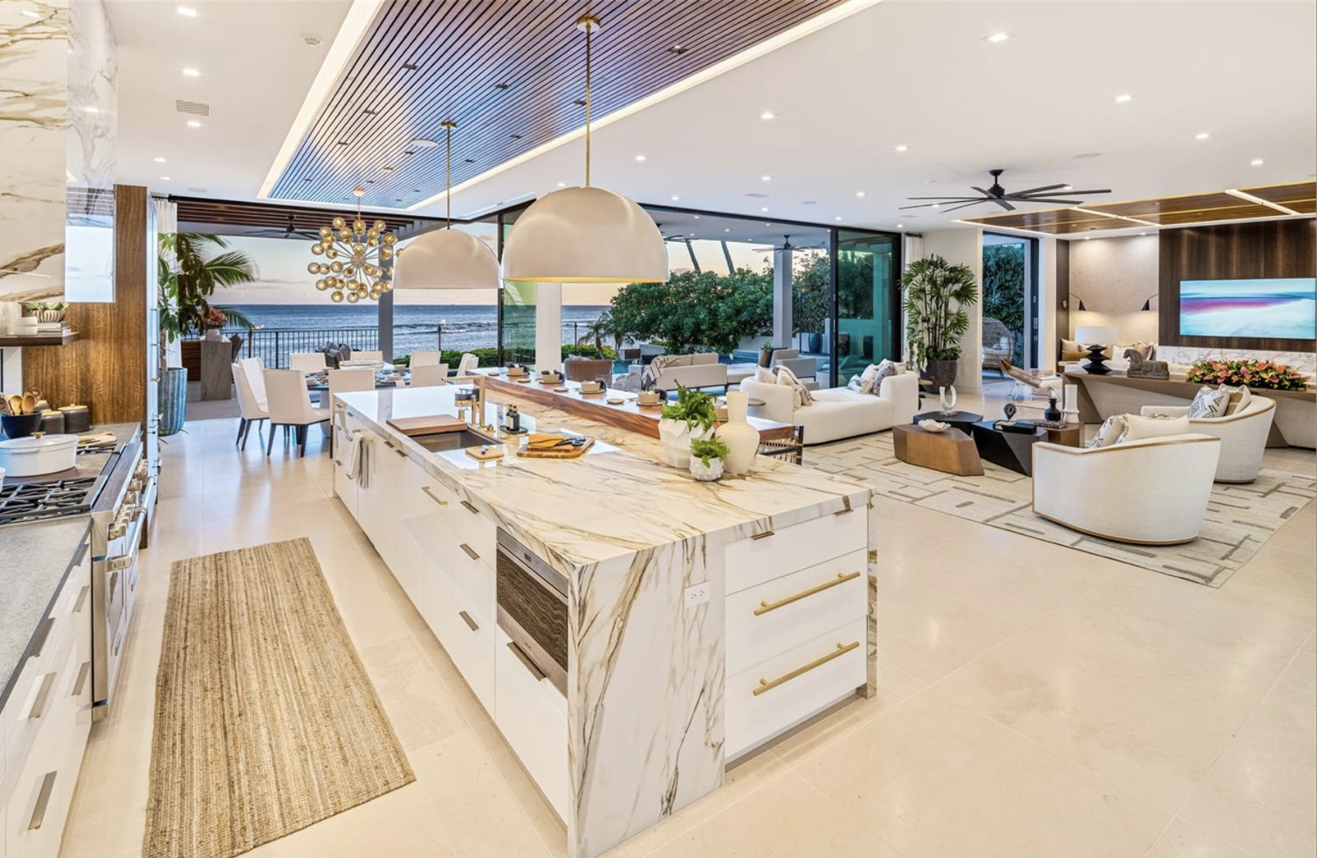 Open-concept living space and kitchen in a modern beach house with ocean view, white furniture, marble island, and large windows.