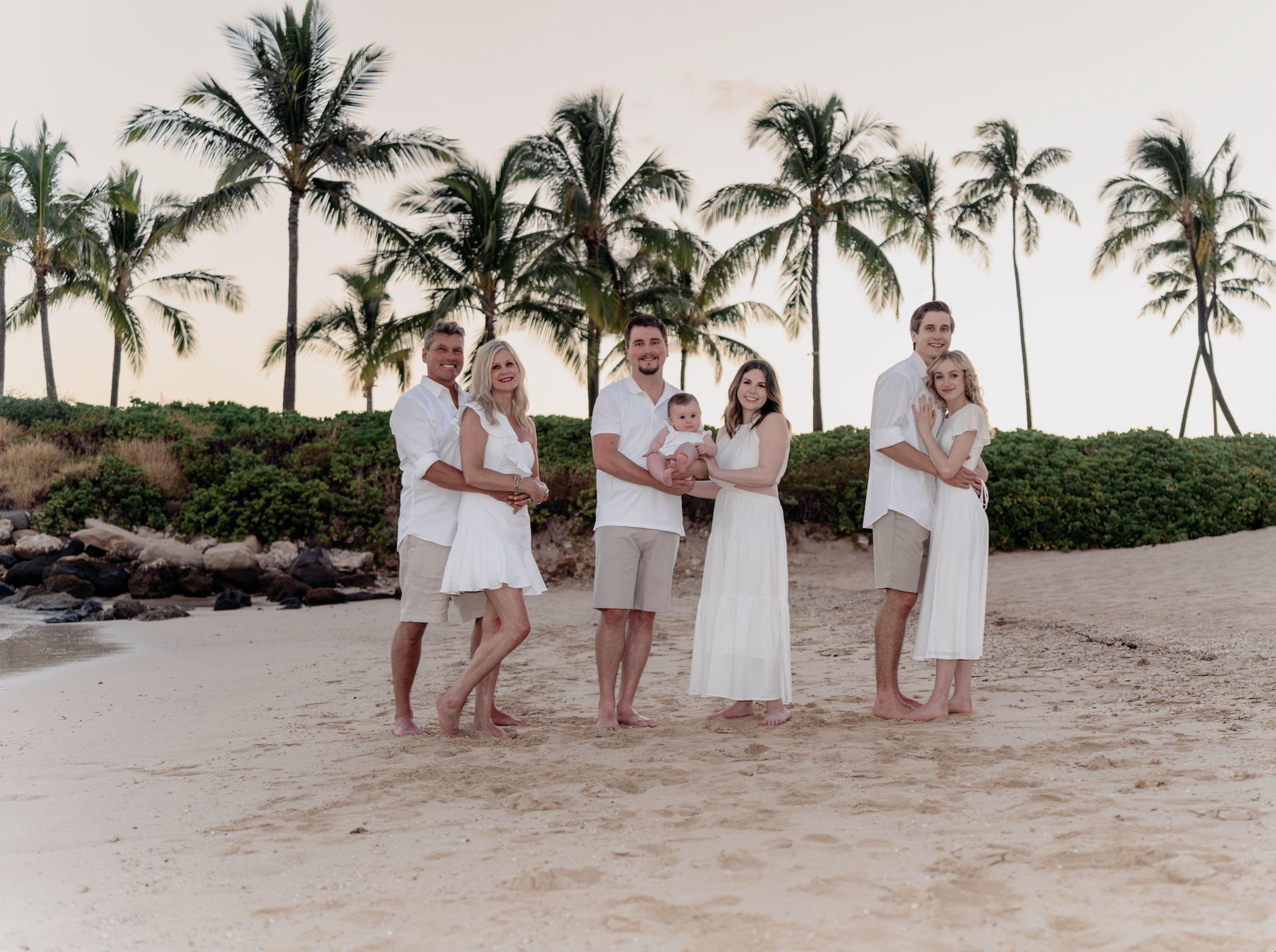 A family of six in white clothing standing on a beach with palm trees in the background during sunset.