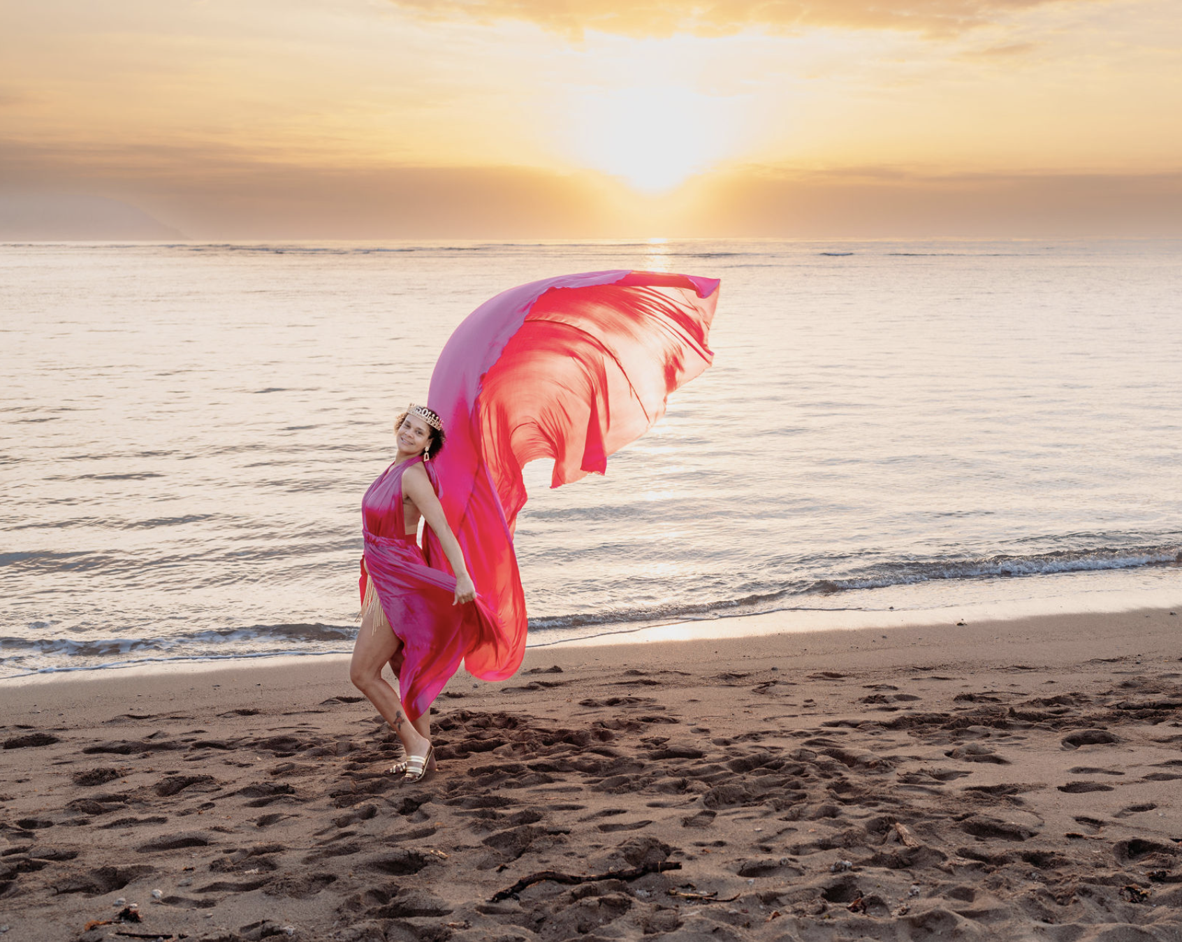 Woman in a pink dress and a crown on her head standing on the beach at sunset, holding a flowing pink fabric.