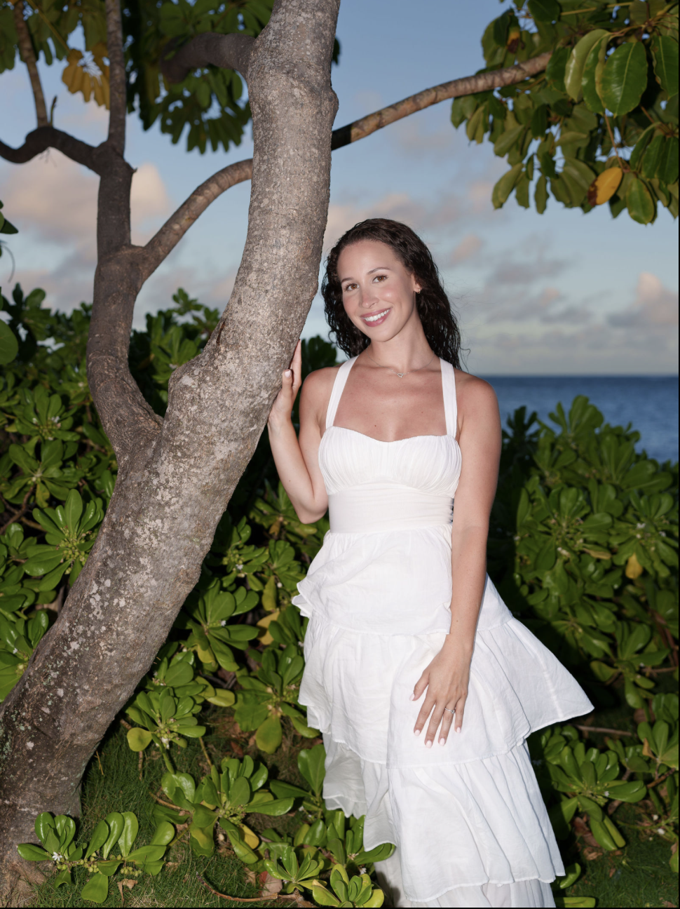 A woman in a white dress standing next to a tree on a lush green tropical landscape with the ocean in the background during sunset.