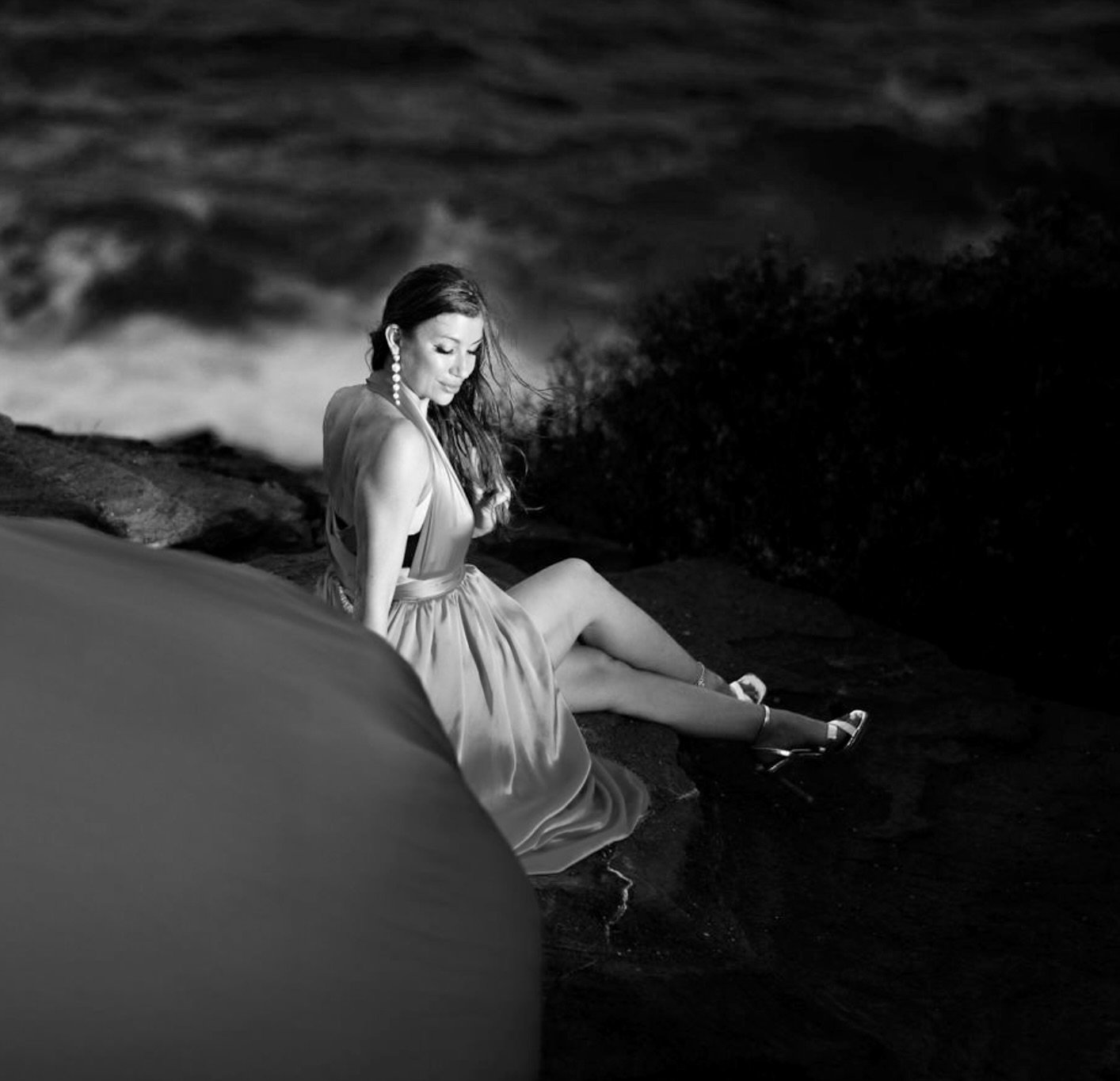 A black-and-white photo of a woman sitting on the rocks near the ocean, wearing a dress and heels, with waves crashing in the background.