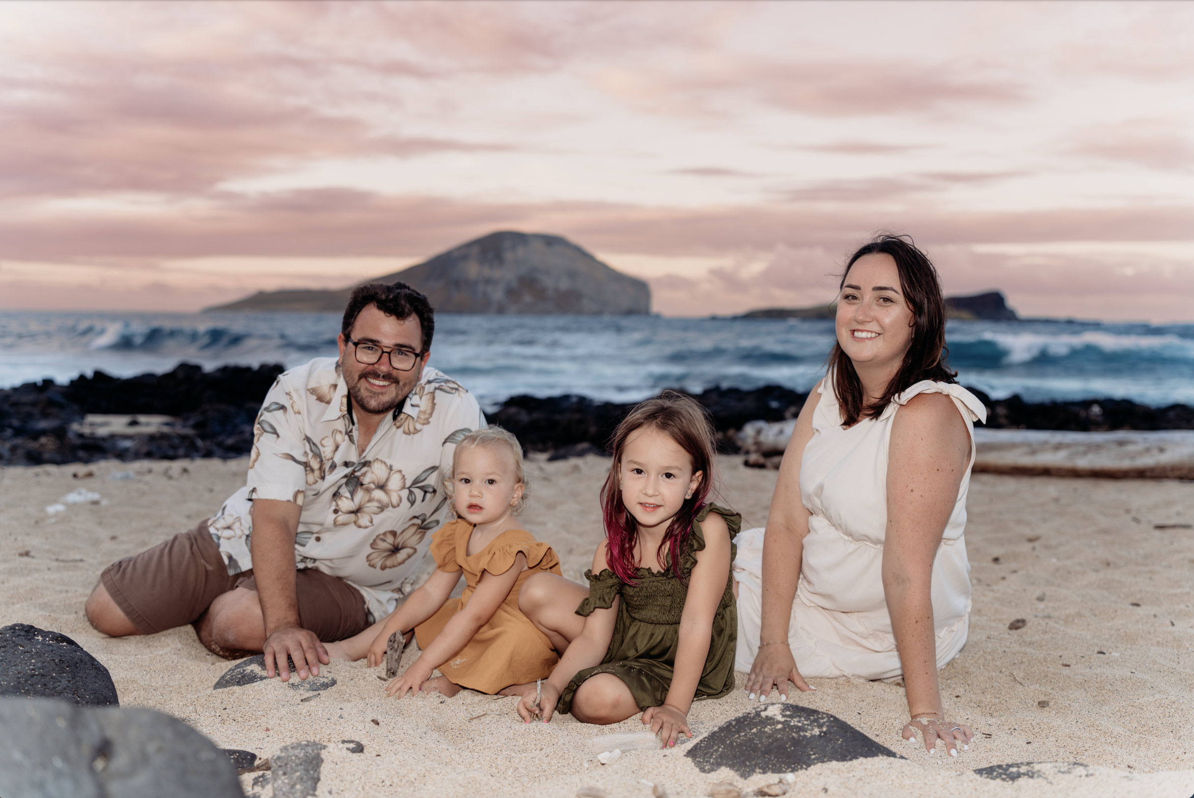 A family of five sitting on a sandy beach with rocks, smiling at the camera, with the ocean and island in the background during sunset.