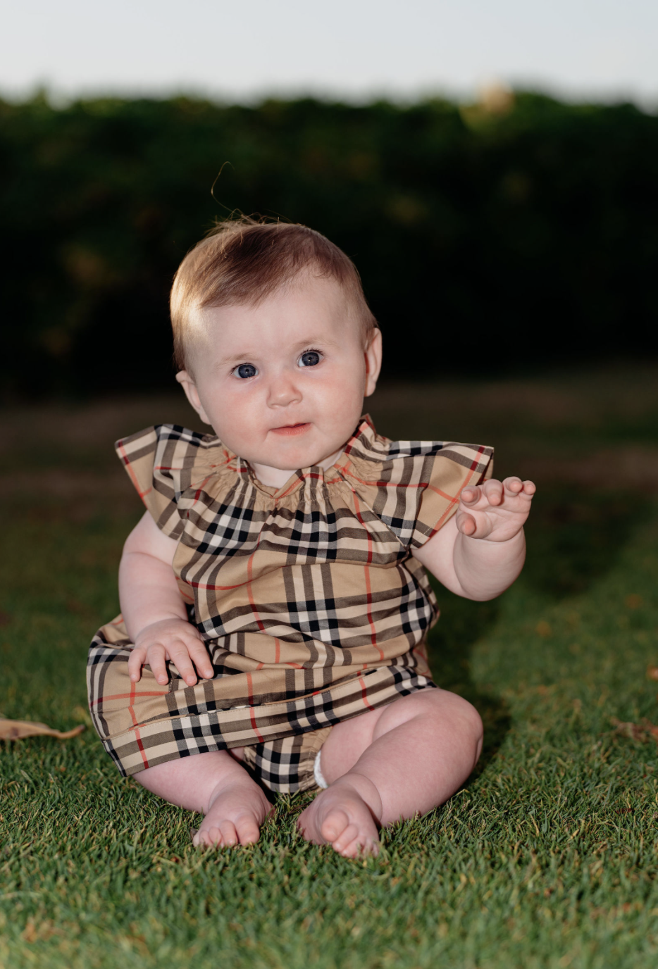 A baby sitting on grass outdoors, wearing a Burberry-patterned dress, with a concerned expression, bushy background, and a small leaf on the ground.