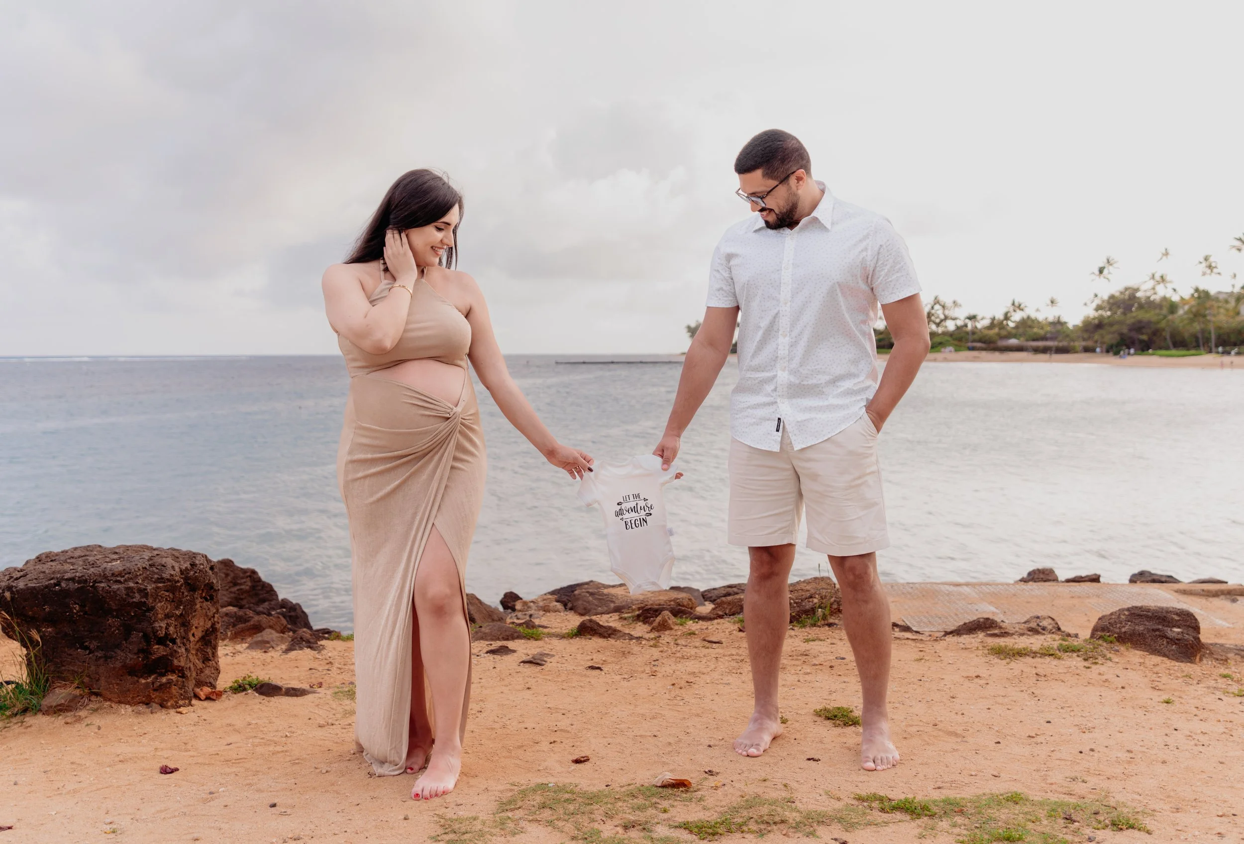 A couple standing barefoot on a beach, holding a small onesie that says 'Let the adventure begin.' The woman wears a beige dress with a slit, and the man wears a white button-up shirt and beige shorts. The ocean and palm trees are in the background.