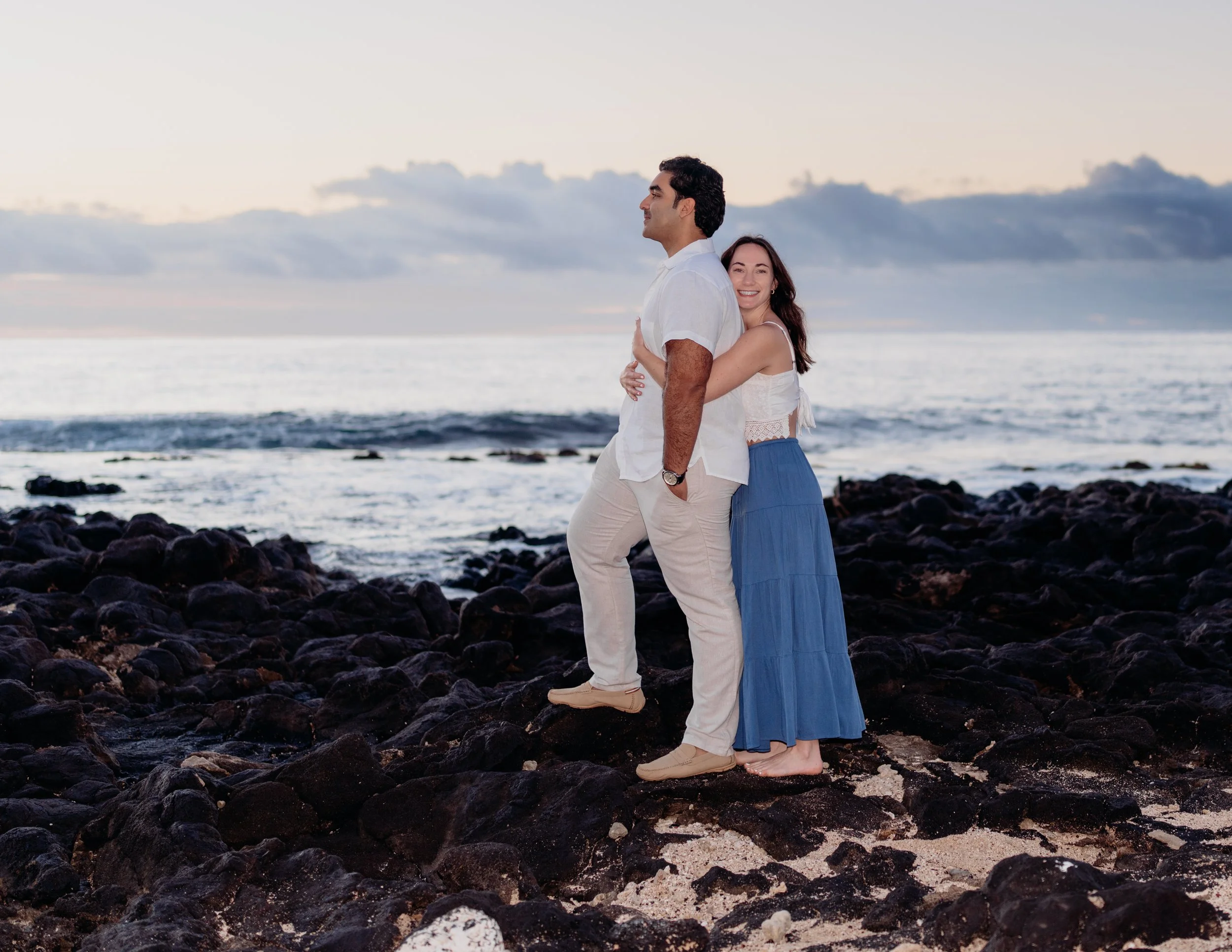 A happy couple stands on a rocky beach at sunset, with the ocean and cloudy sky in the background. The woman embraces the man from behind, both smiling.
